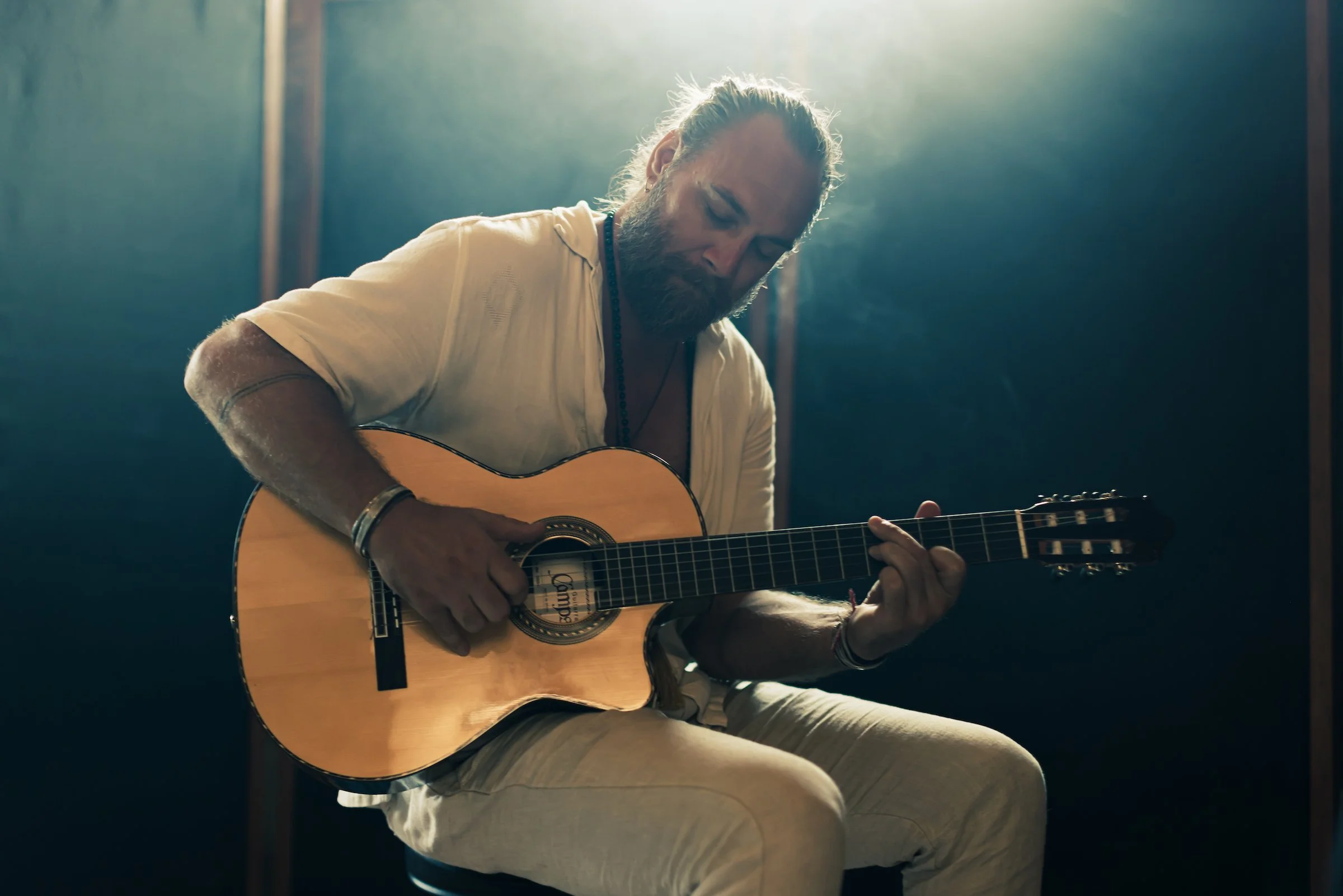 A man with a beard and long hair in a bun, wearing a light-colored shirt and pants, playing an acoustic guitar in a dimly lit room.