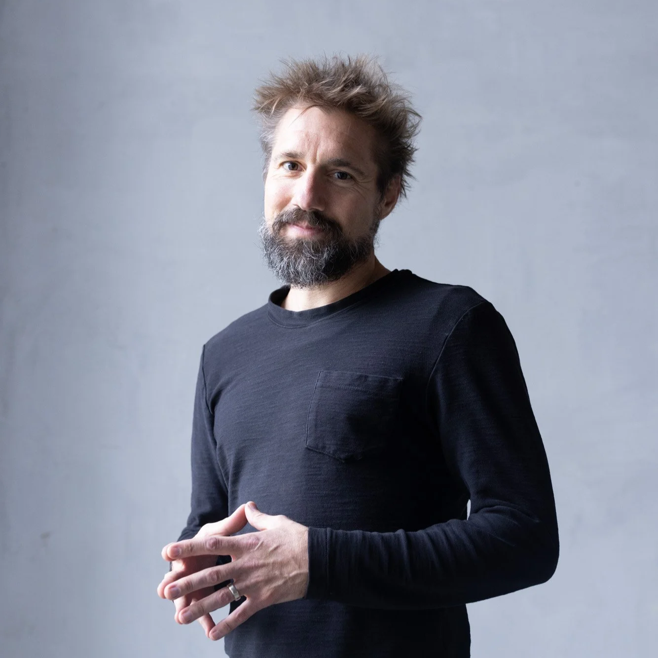 Ty Landrum, yoga teacher dressed in black with his hands touching each other, smiling, with a grey back ground