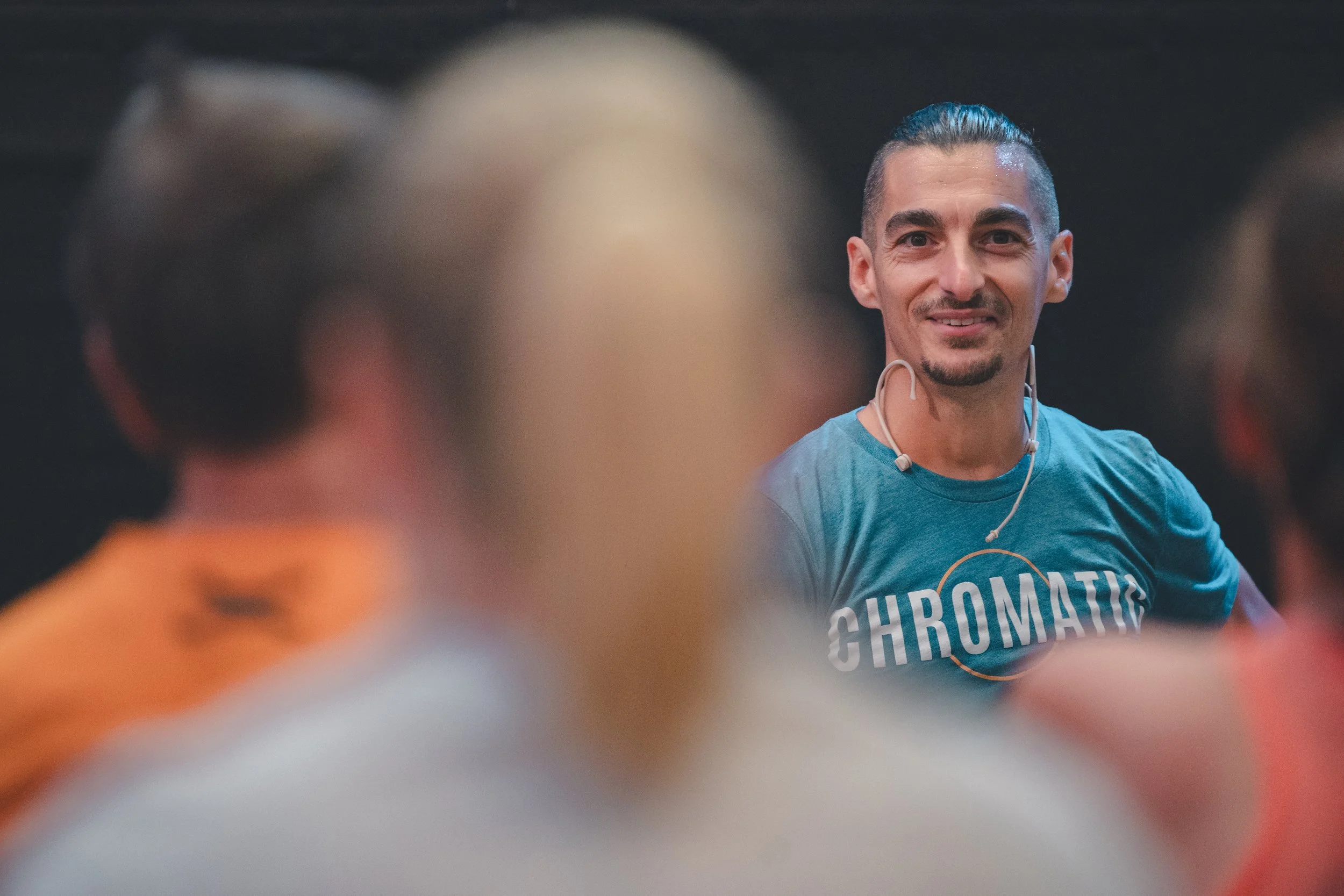 A smiling male instructor with dark hair tied back, wearing a blue shirt with the word 'CHROMATIC' and a headset, speaking to a group of people in a classroom or seminar setting.
