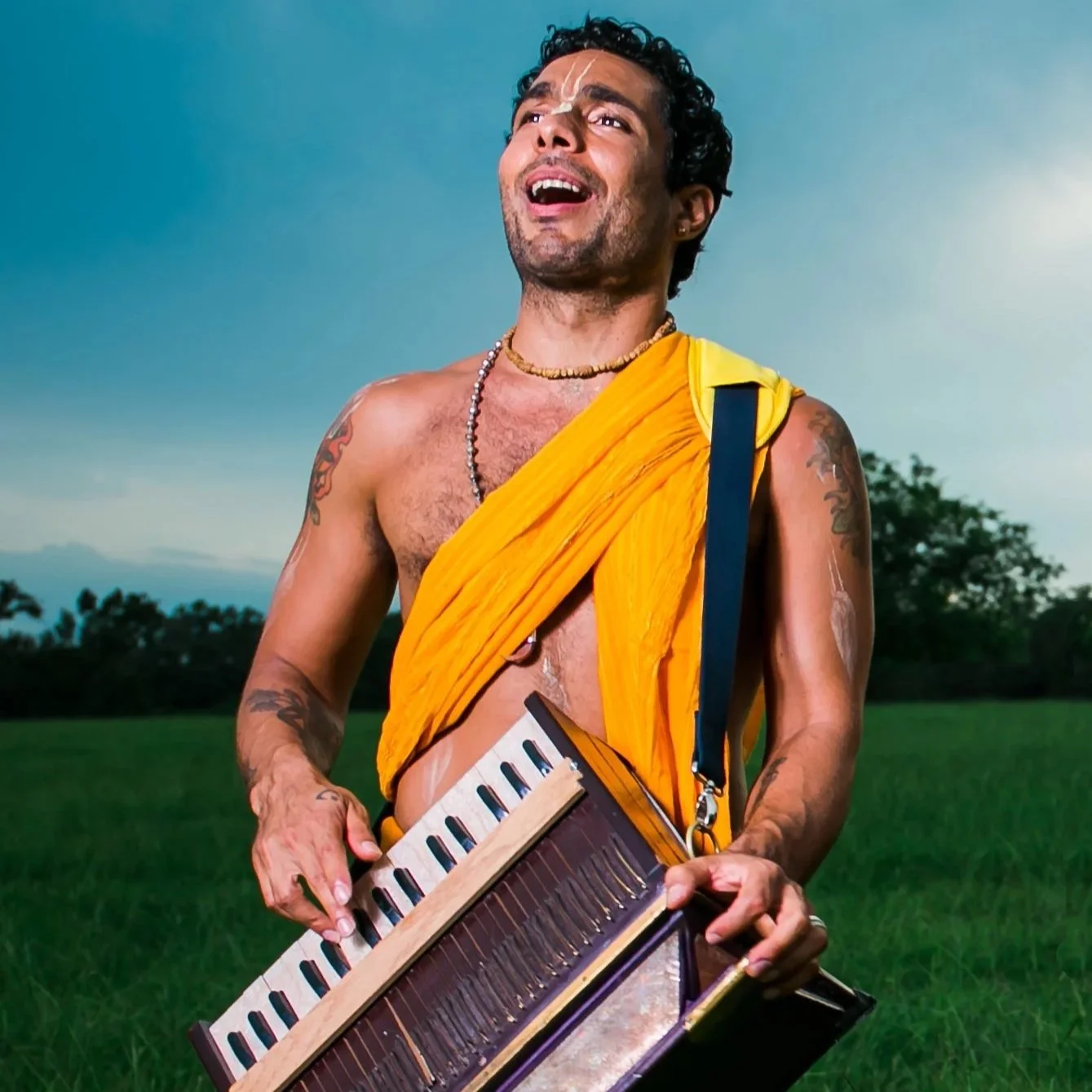 A man with tattoos wearing a yellow saffron cloth and necklaces, holding a harmonium outdoors with greenery and a blue sky in the background.