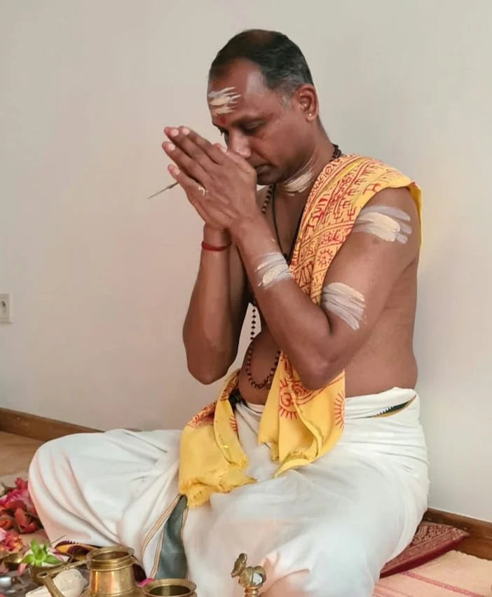 A Hindu priest in traditional attire praying with hands pressed together, seated on the floor with ritual items nearby.