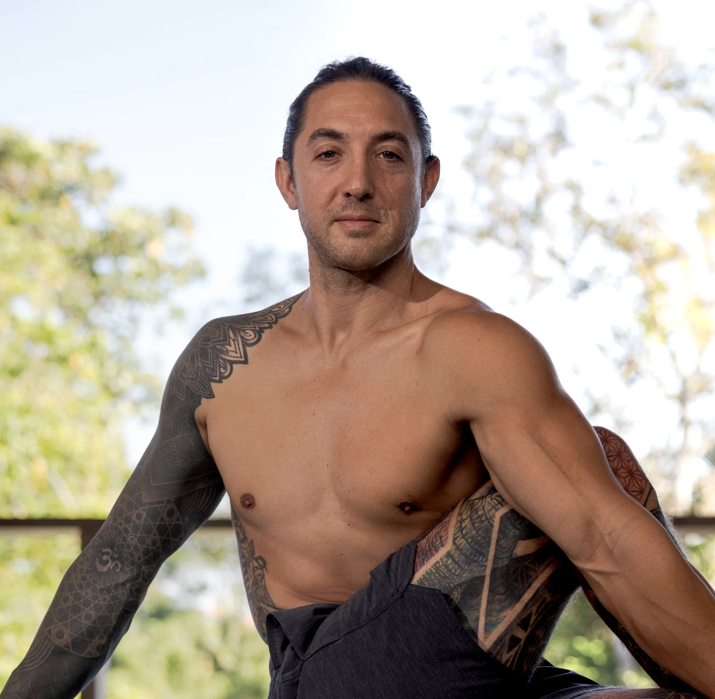 Man with tattoos and a naked upper body sitting in a twist looking in the camera