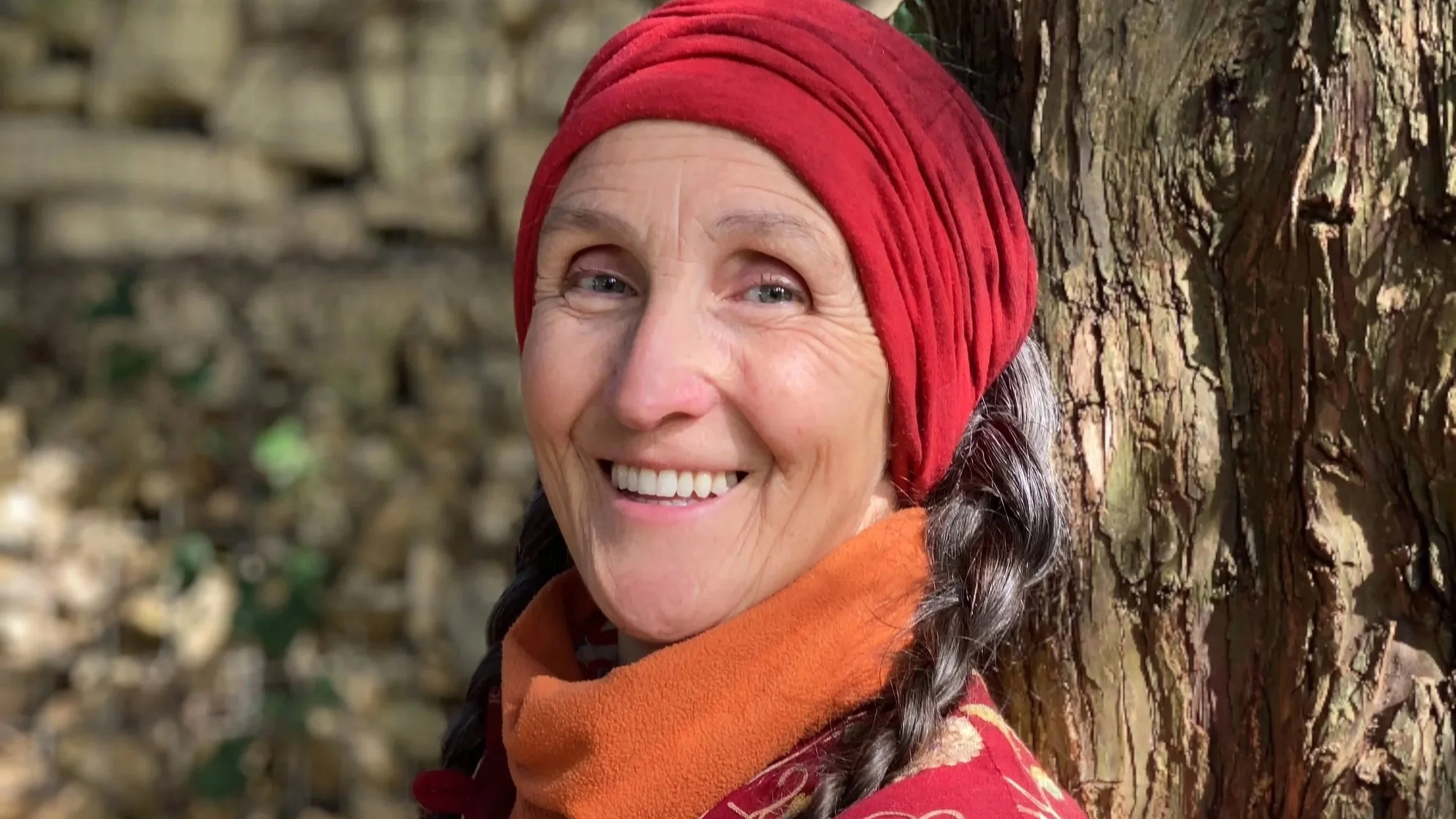 Close-up of a smiling woman with gray hair in braids, wearing a red headscarf and orange scarf, leaning against a tree in an outdoor setting.