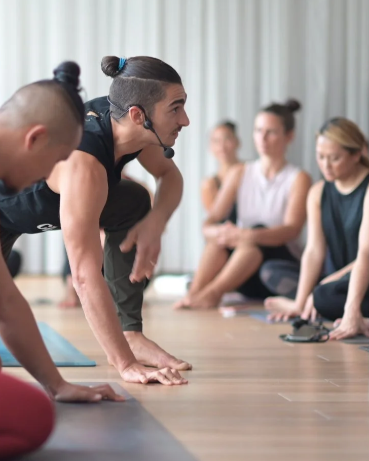 Instructor leading a yoga class in a studio with participants sitting and observing.