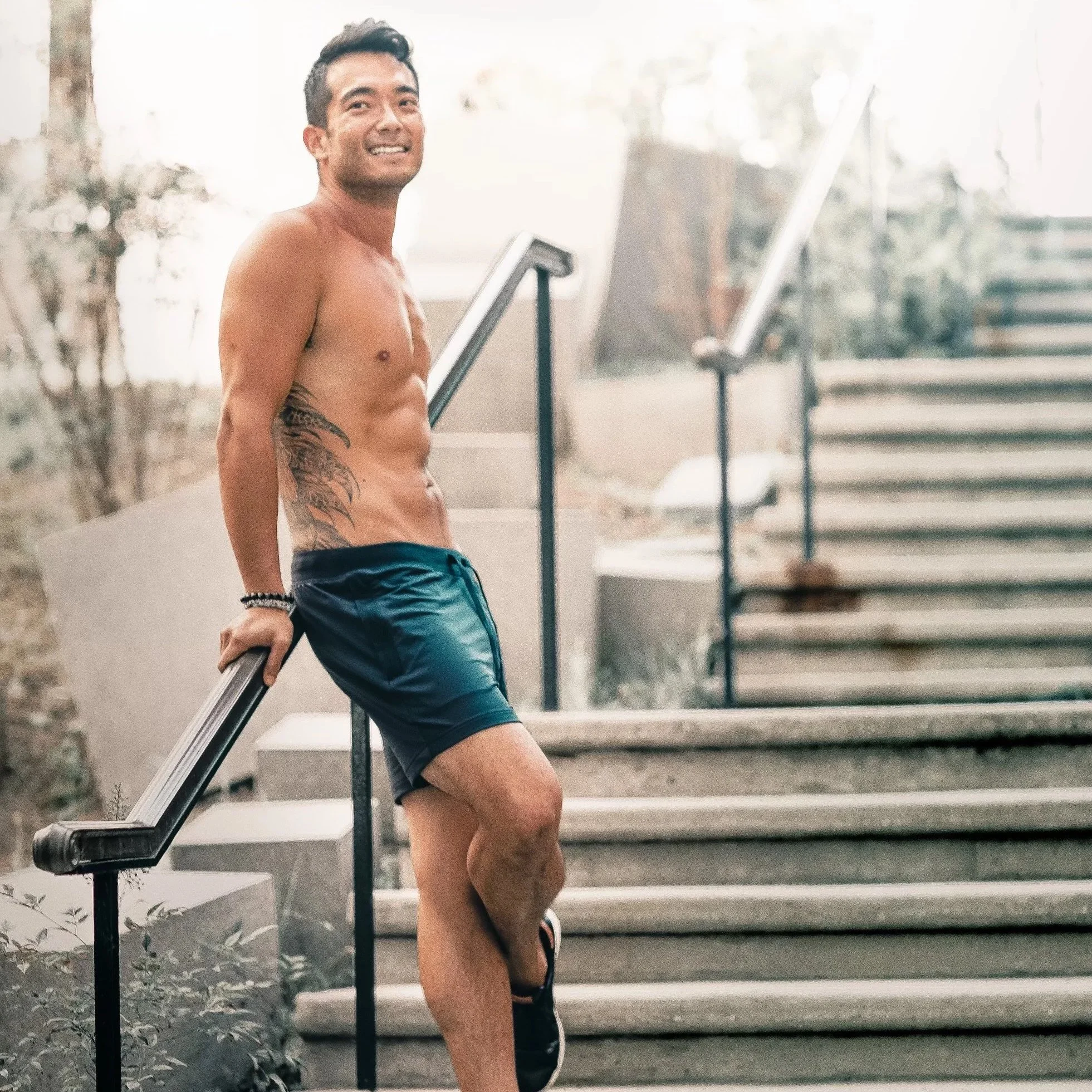 Man standing outside on concrete stairs leaning against the rail wearing only black shorts