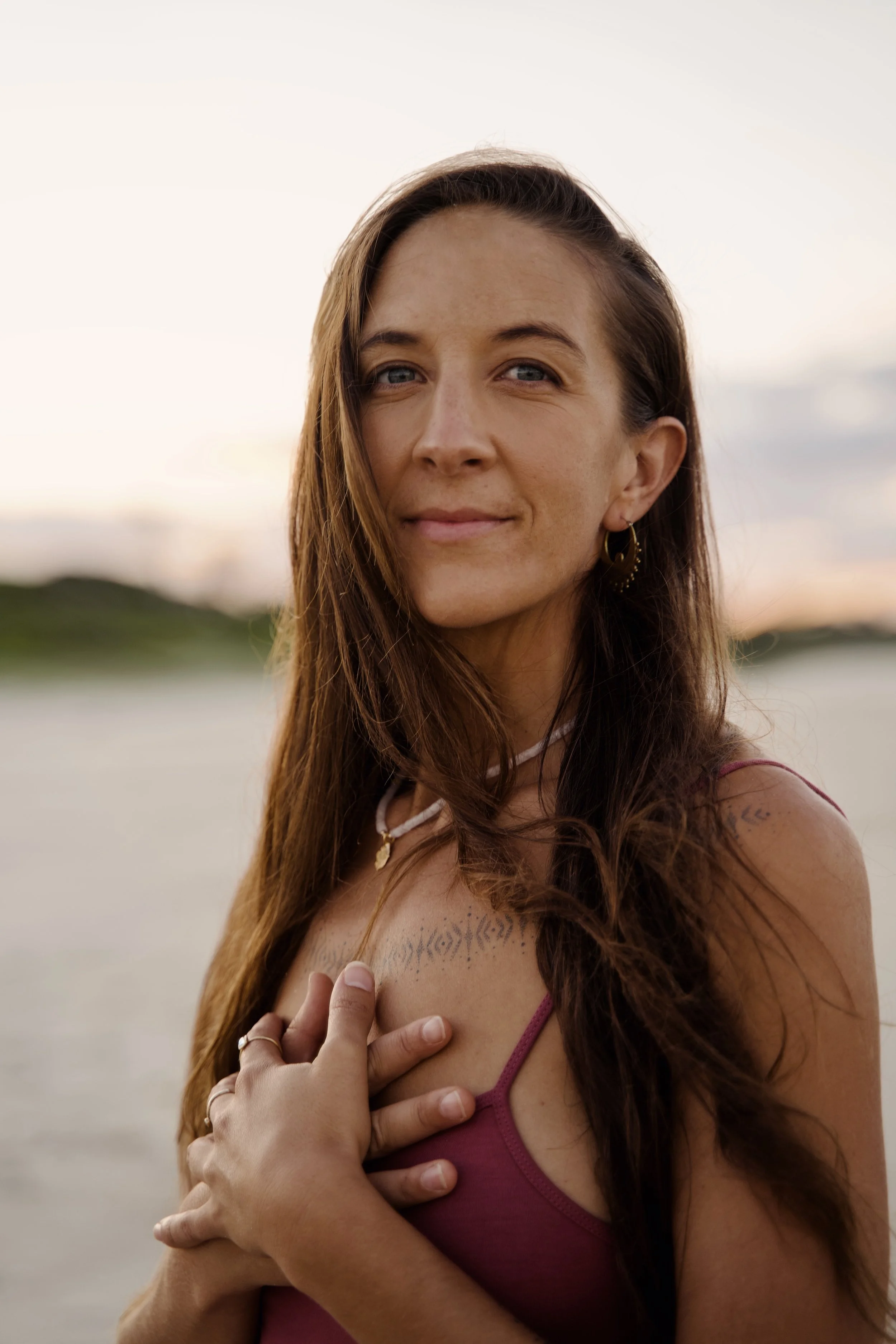 A woman with long brown hair and blue eyes, wearing a maroon tank top, standing outdoors at sunset near water, with her hand on her chest and a tattoo visible on her collarbone.