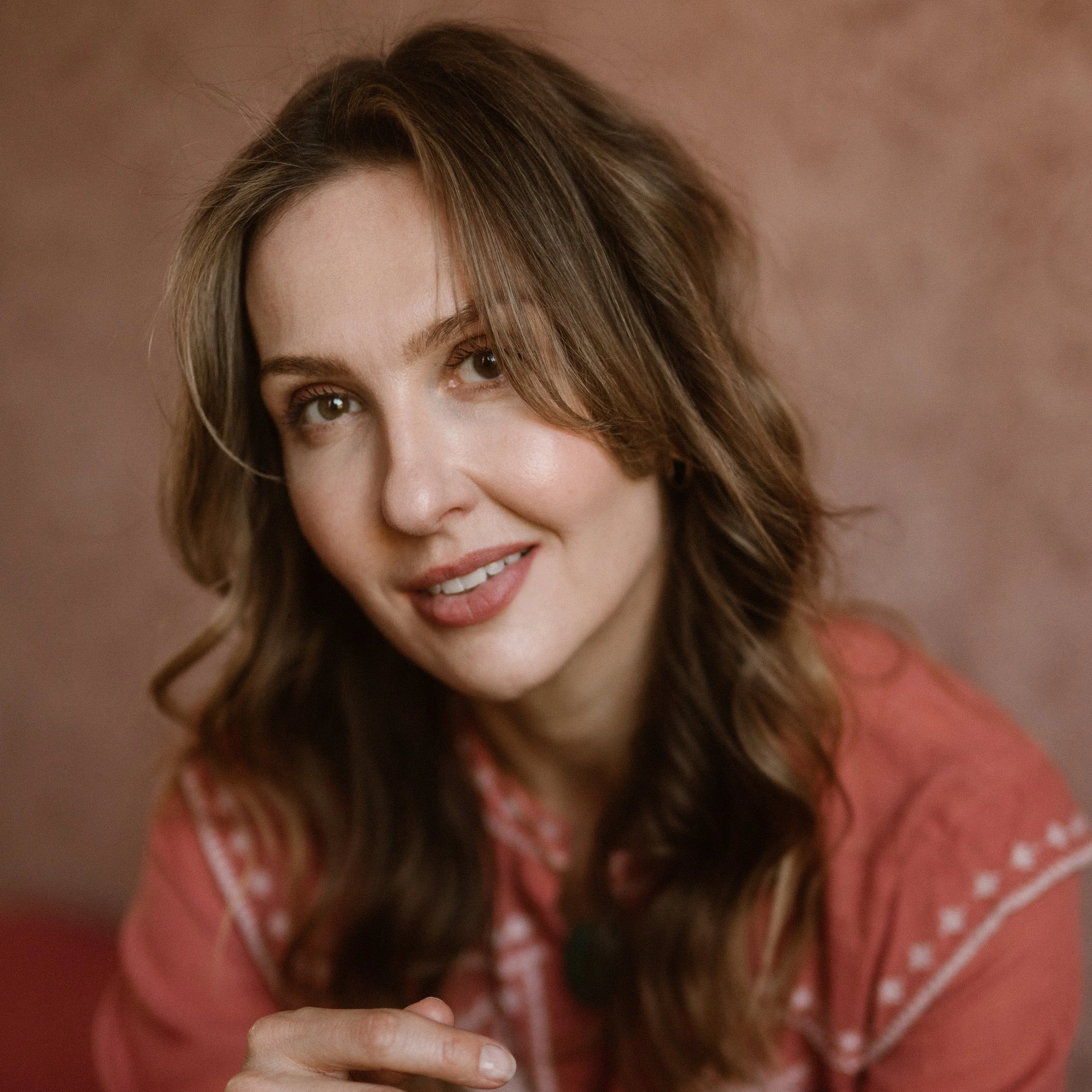 Close-up of a smiling woman with shoulder-length brown hair, wearing a red top, against a muted pink background.