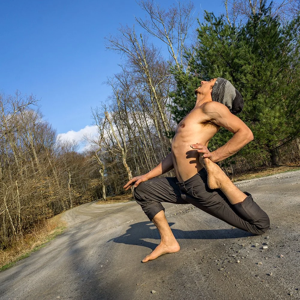 A shirtless man with a head covering performs a yoga pose on a gravel road surrounded by leafless trees and green bushes under a clear blue sky.