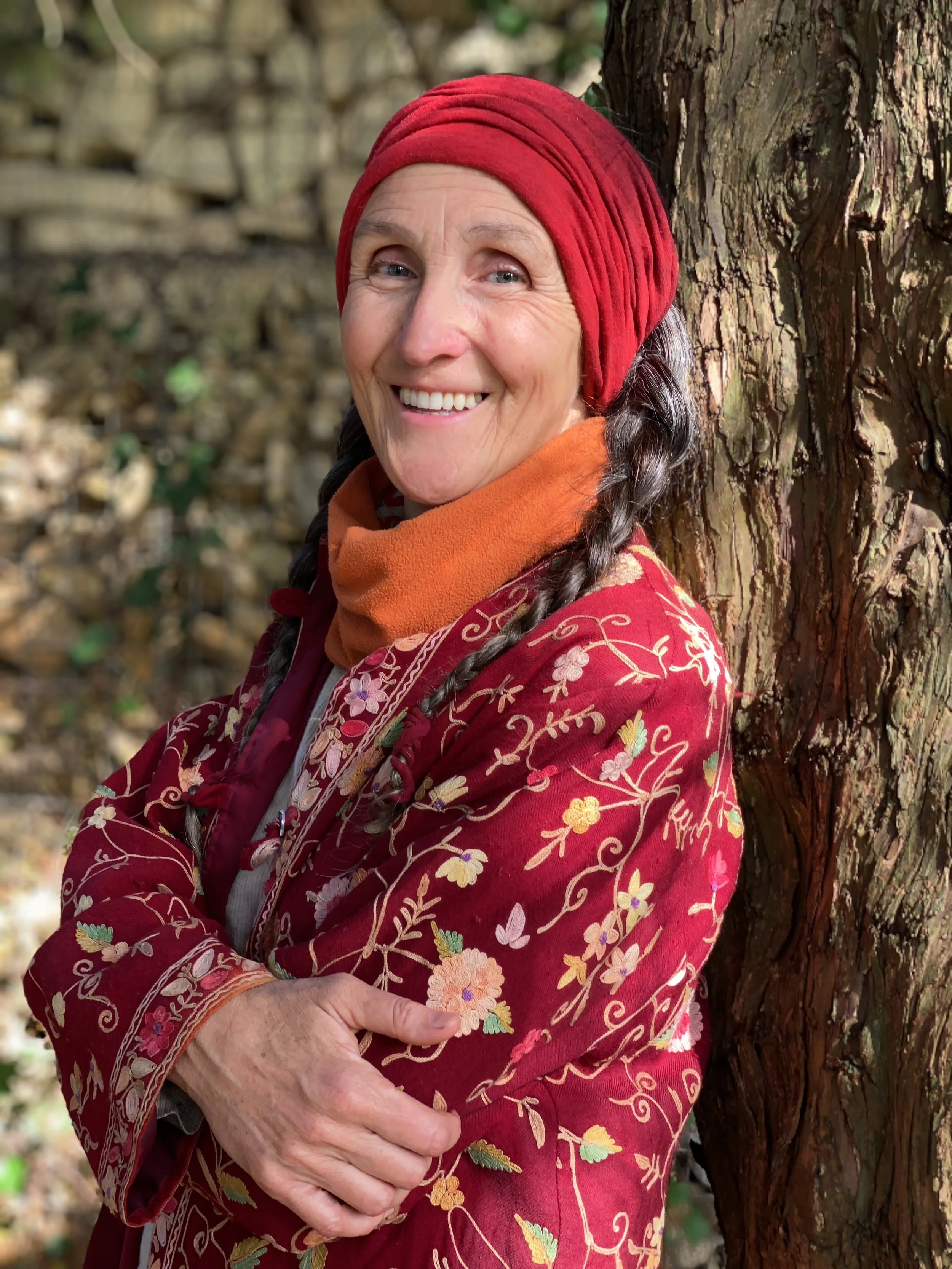 A woman in a red floral embroidered coat, orange scarf, and red headwrap standing outdoors next to a tree, smiling at the camera.