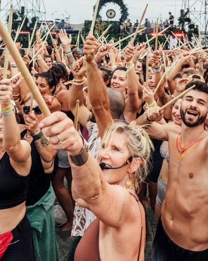 Crowd of people at an outdoor music festival, with many holding drumsticks, dancing, and smiling, under a cloudy sky.