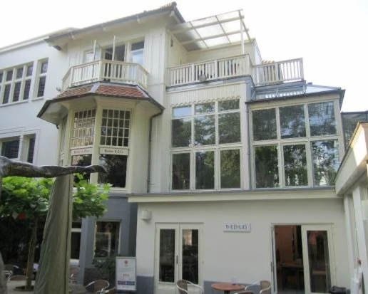 A multi-story white building with large windows, small balconies, and outdoor seating area on the ground level.