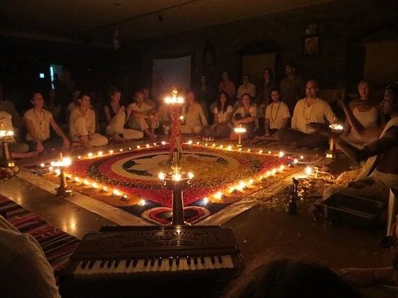 People gathered in a circle around a large decorated floor of flowers, with lit candles and oil lamps, in a dimly lit room, celebrating a traditional festival.