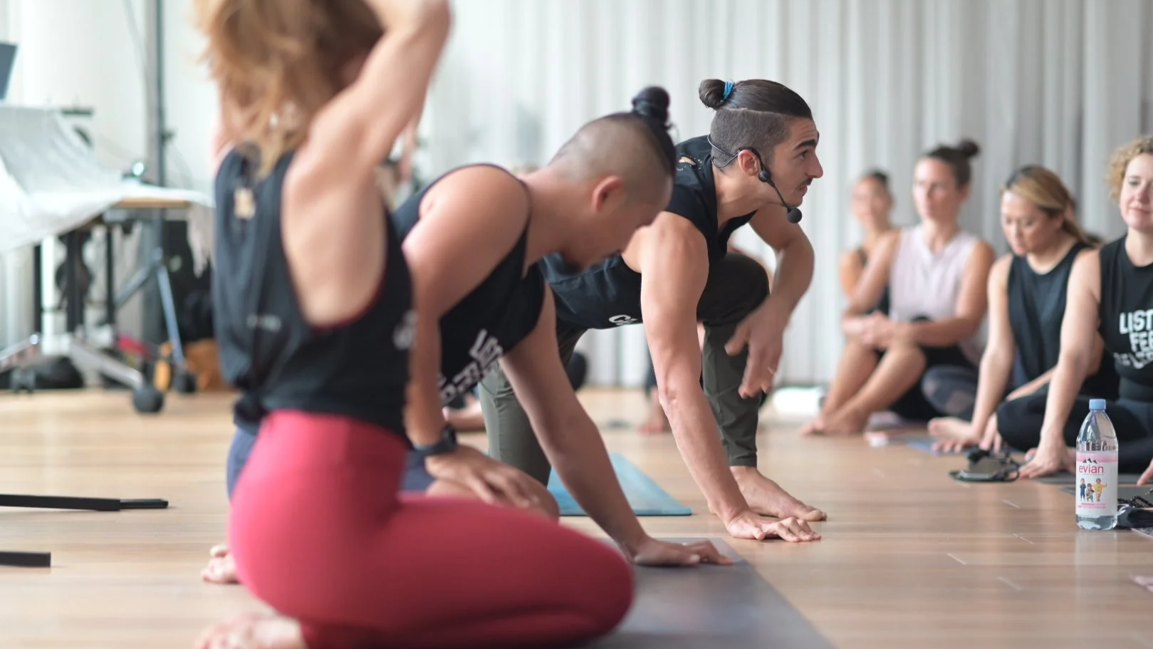 Yoga instructor in black tank top demonstrates yoga pose on wooden floor, surrounded by women seated in yoga class, with water bottle nearby.