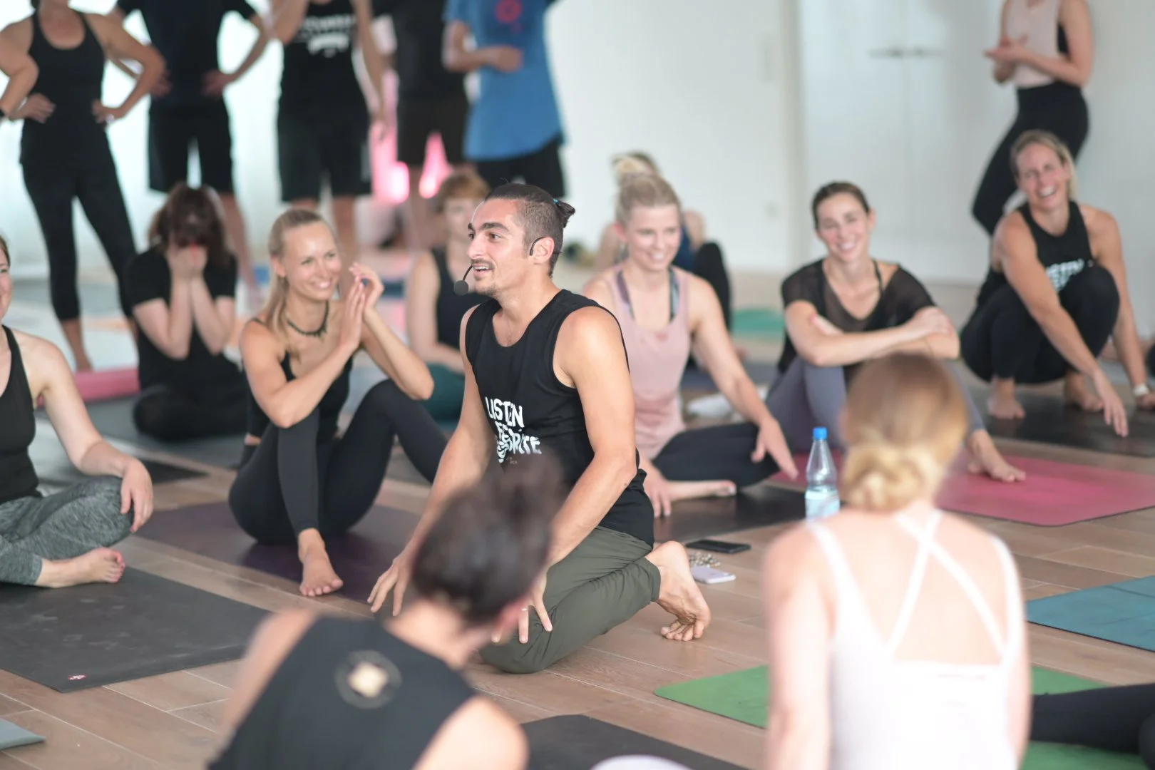 A group of people participating in a yoga class, with a male instructor leading the session while sitting on his knees, surrounded by women sitting on yoga mats in a bright room.