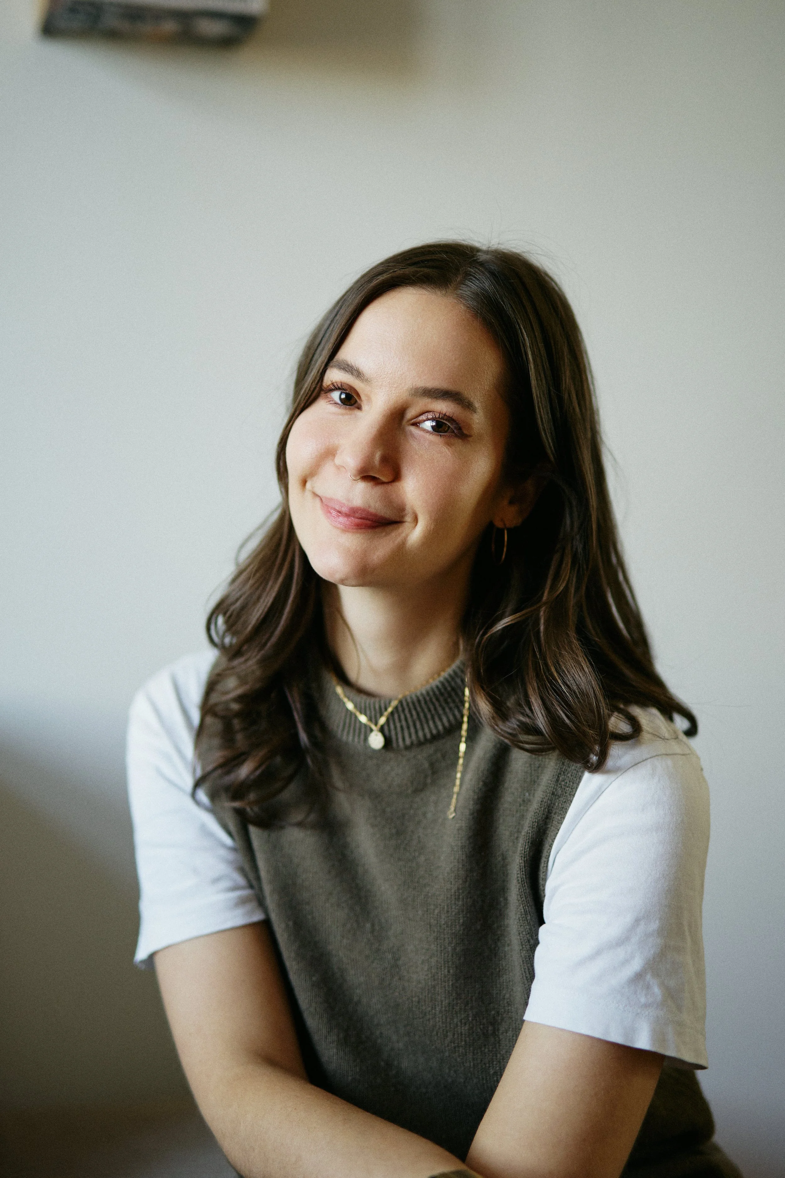 Porträt einer jungen Frau mit braunen, lockigen Haaren, die lächelnd in die Kamera schaut, vor einem neutralen Hintergrund.
