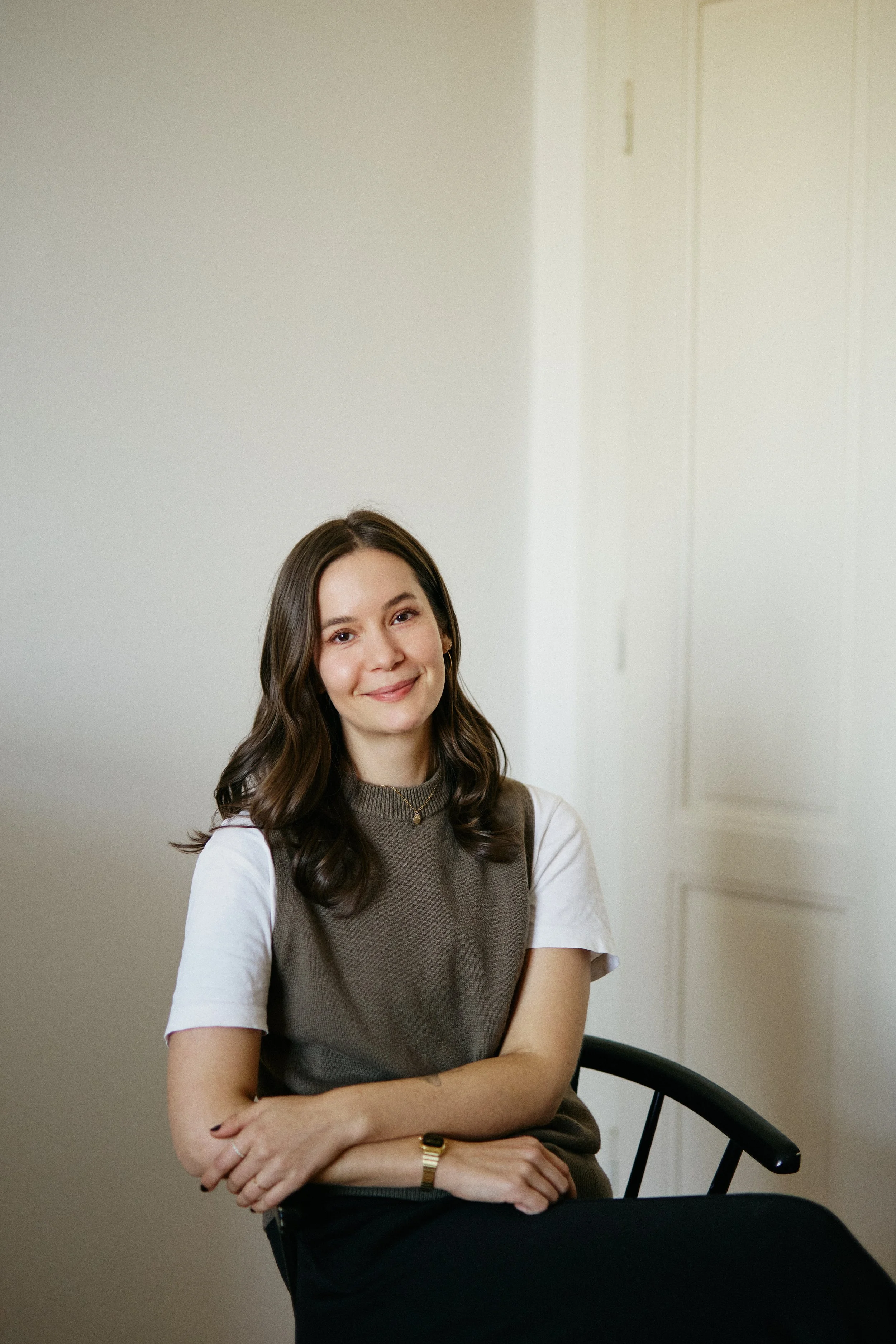 Ein lächelnde junge Frau mit braunen, welligen Haaren sitzt in einem Raum mit weißen Wänden, trägt ein graues Oberteil mit weißen T-Shirts darunter, eine Armbanduhr, und hat ihre Arme verschränkt.