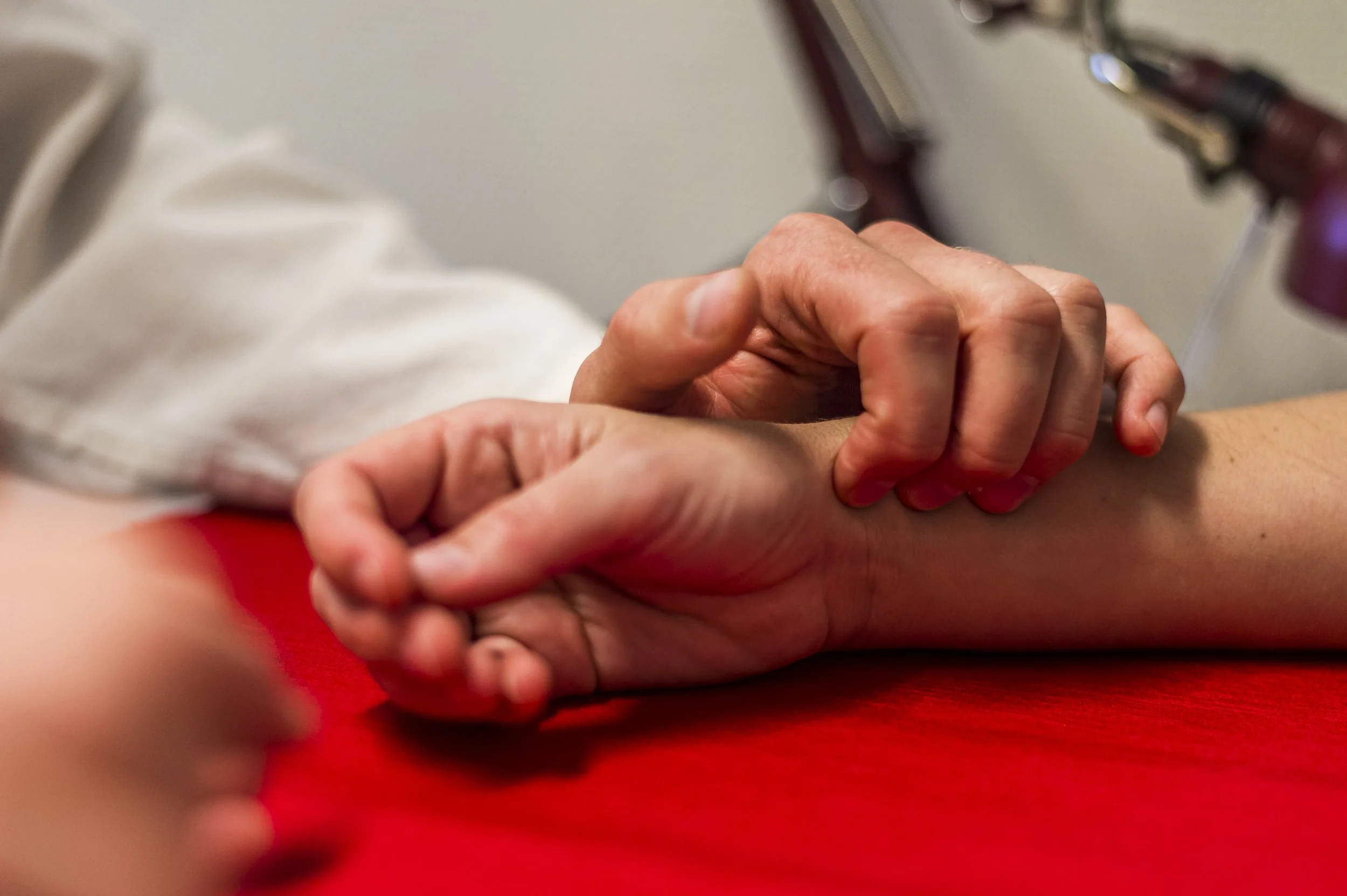 Närbild på en person som håller en annan persons hand, vilket kan indikera ett uttryck för vård eller tröst, med en röd matta och medicinsk utrustning i bakgrunden.