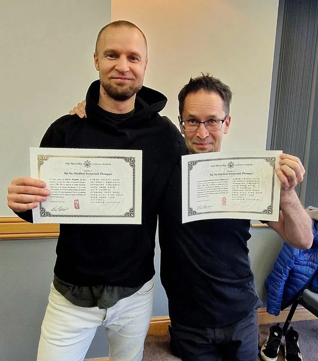 Två män håller upp diplom, står i ett rum med beige vägg och en träningsjacka på en stol i bakgrunden.