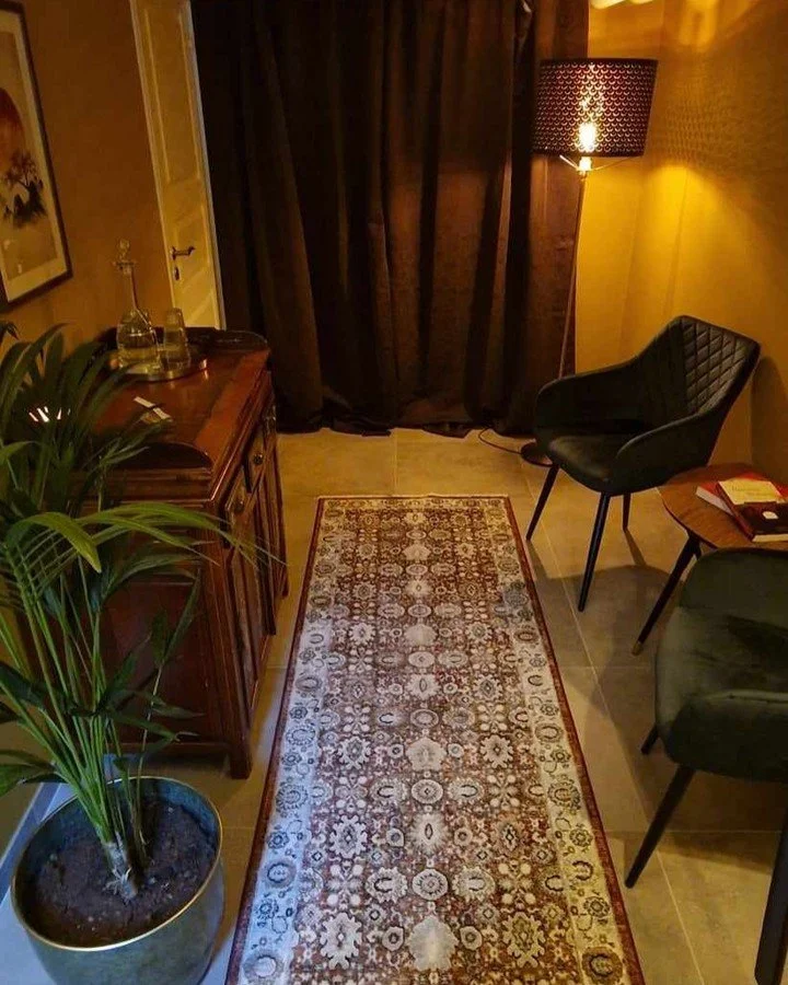 Ett mysigt kontorsutrymme med en matta, fåtöljer, ett sidobord med böcker, en växt och en bordsd840;gardiner, samt en golvlampa som ger ett varmt ljus.