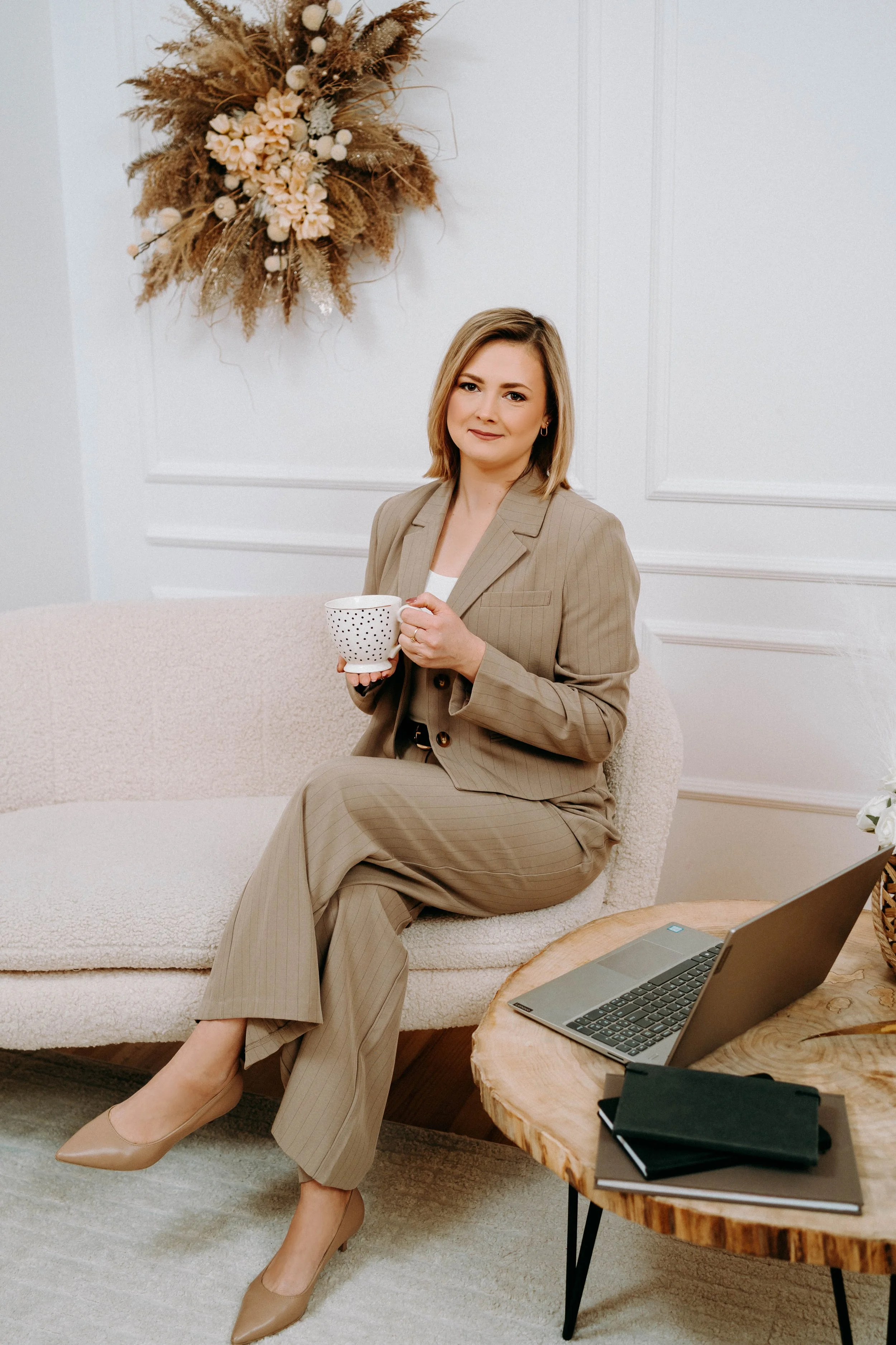A woman in a beige pantsuit sitting on a light-colored sofa, holding a white mug with black polka dots, with a laptop and notebooks on a wooden table beside her, and a beige floral wreath hanging on the white wall behind her.