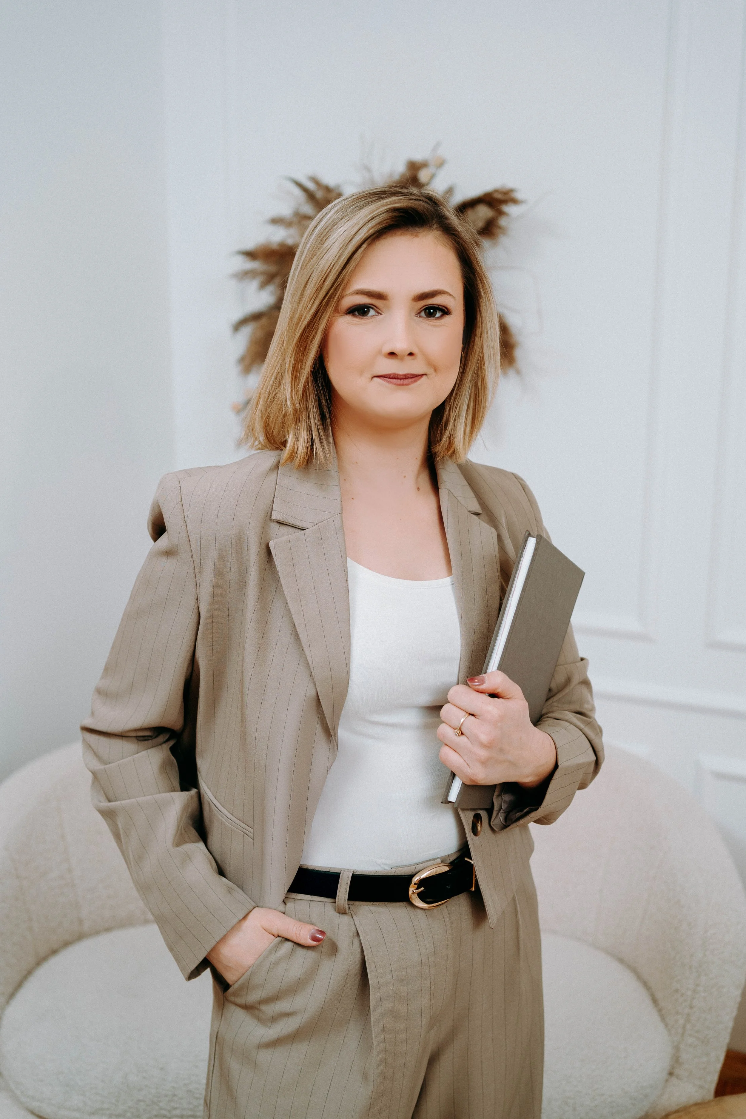 A woman with shoulder-length blonde hair wearing a beige pinstripe suit and white shirt, holding a notebook, standing indoors in front of a white wall with a decorative arrangement of dried plants.