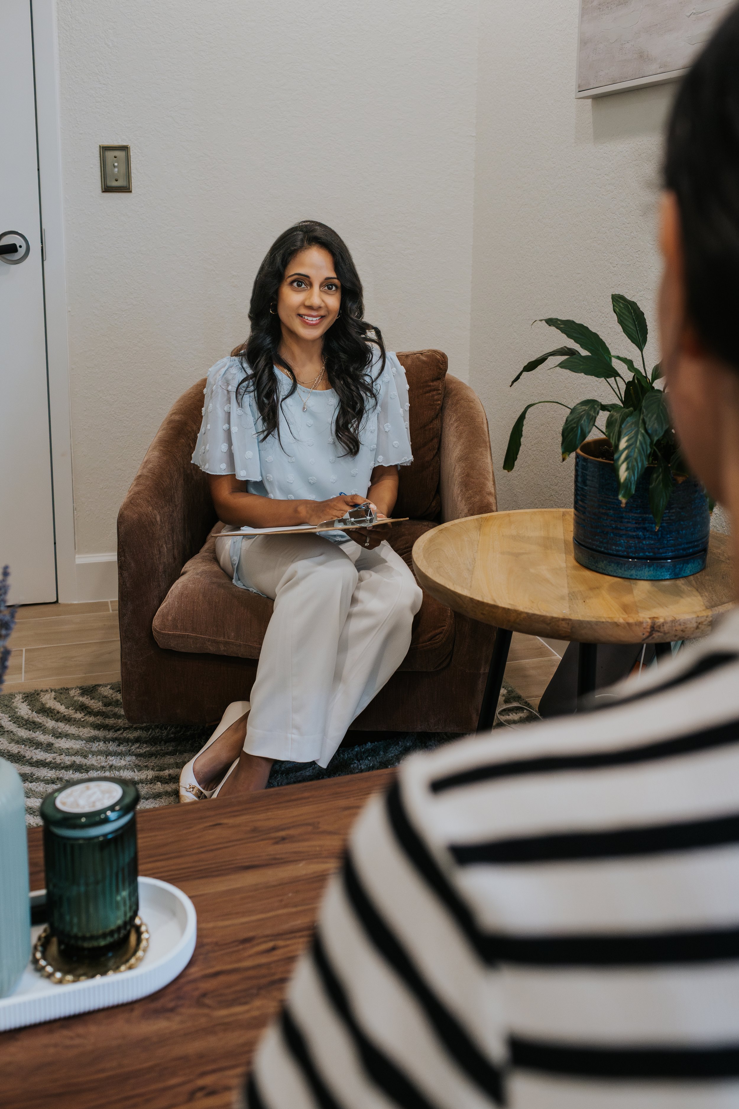 A woman with long dark hair sitting on a brown couch with a clipboard in her hand, smiling and facing a person in a black and white striped shirt in a room with a plant on a round wooden table.