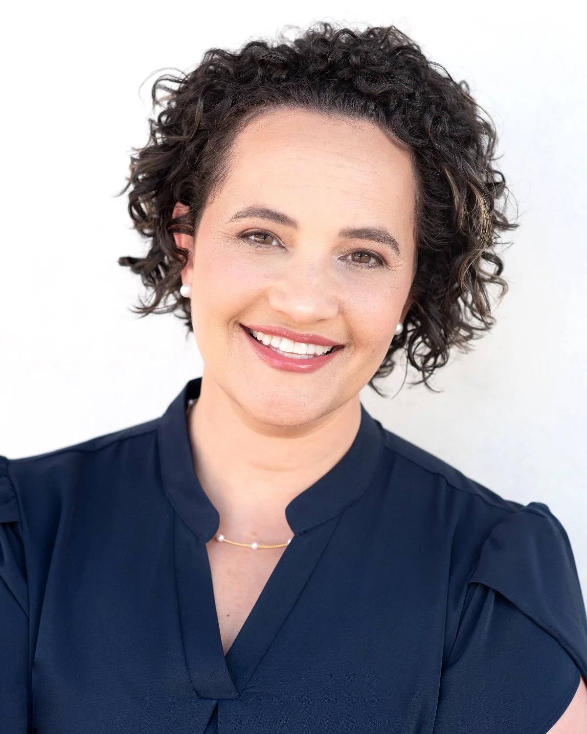 Portrait of a smiling woman with short, curly dark hair, wearing pearl earrings, a pearl necklace, and a navy blouse against a plain white background.