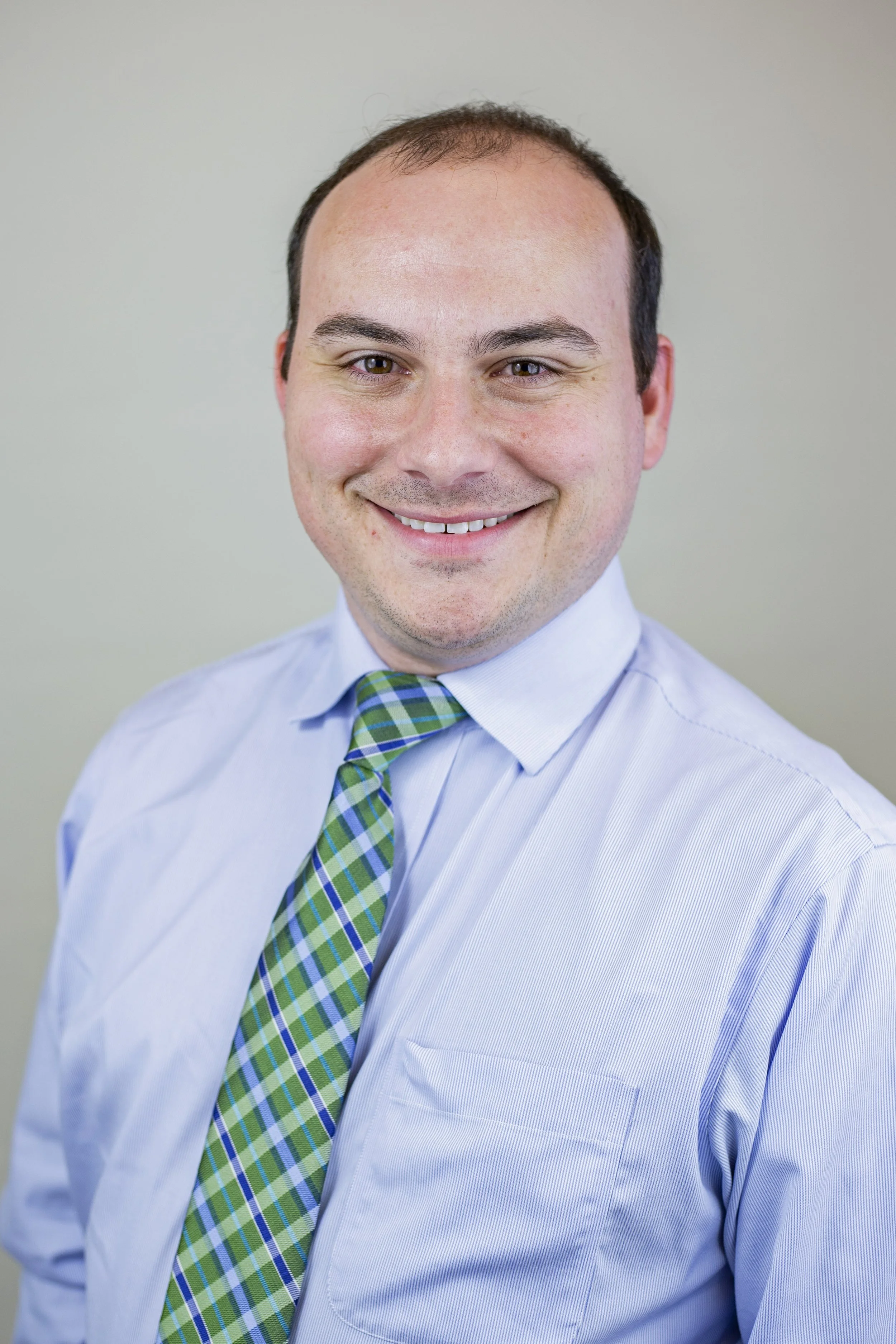 Headshot of a young man with short dark hair, smiling, wearing a light blue dress shirt and a green plaid tie.