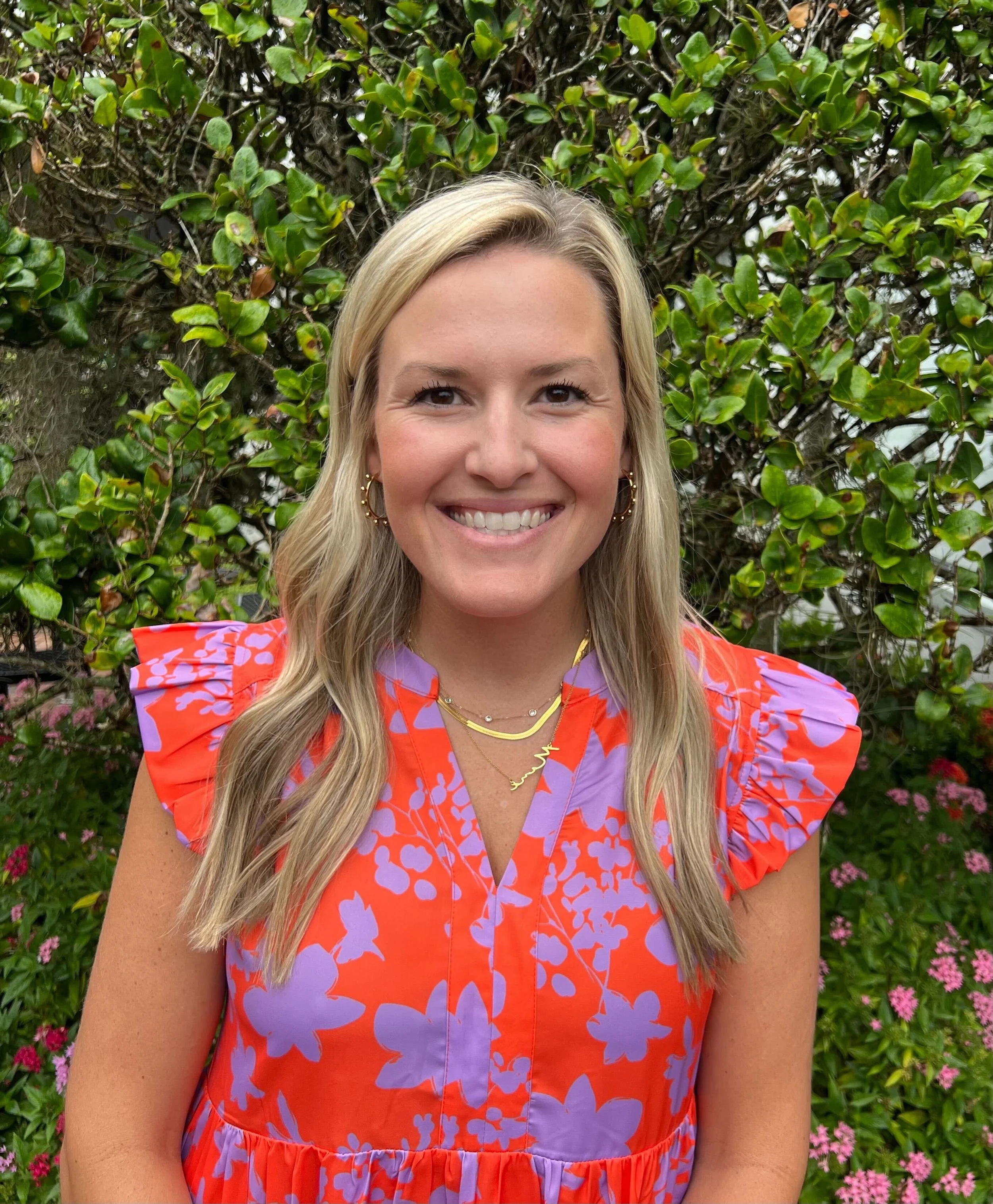 Close-up of a smiling woman with blonde hair, wearing gold jewelry, in front of a green leafy background with pink flowers.