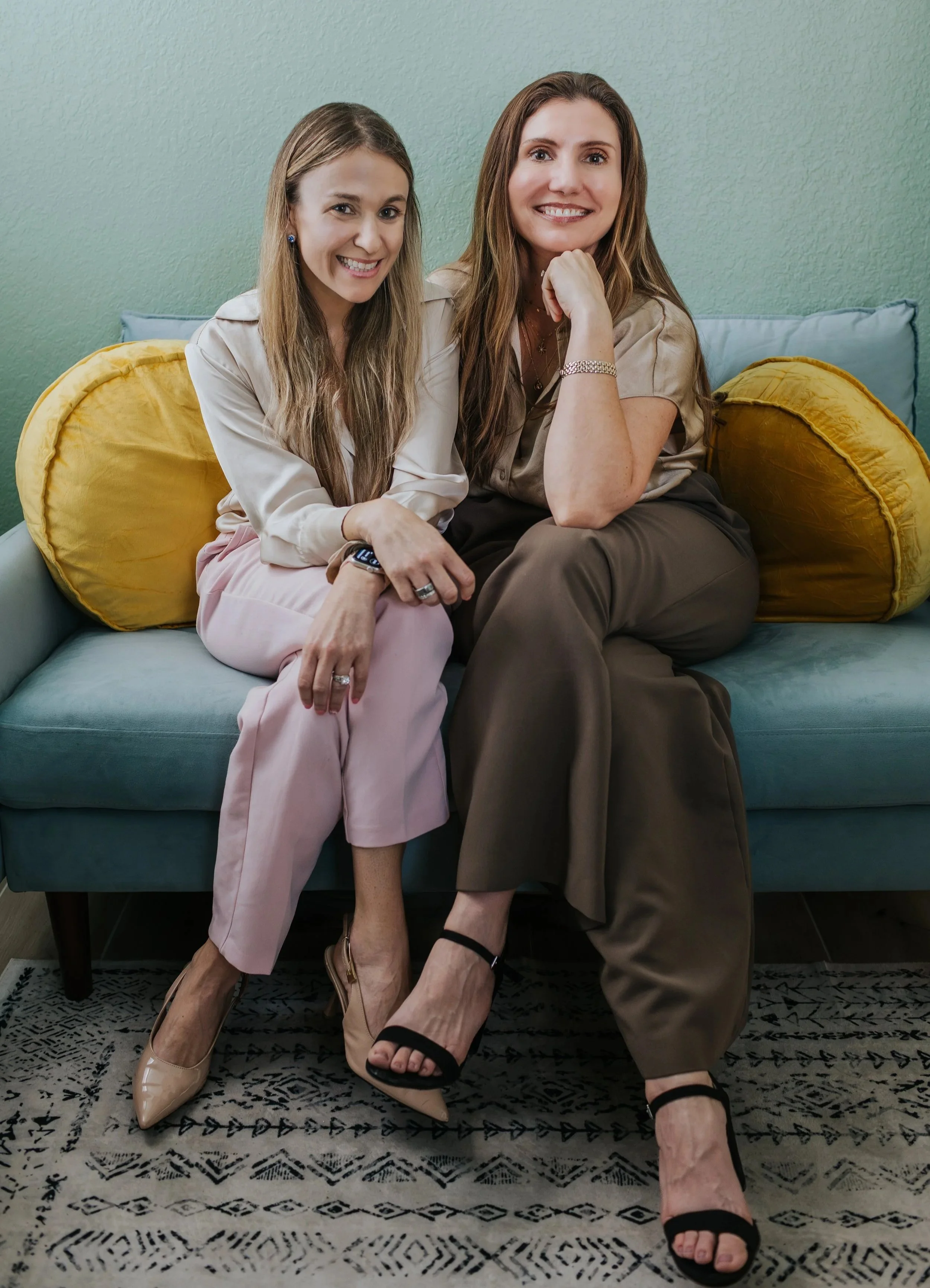 Two women sitting on a blue sofa with yellow pillows, smiling, one with long blonde hair and light pink pants, the other with long brown hair and brown pants, both in beige tops, wearing high heels, in a room with a green wall and patterned rug.