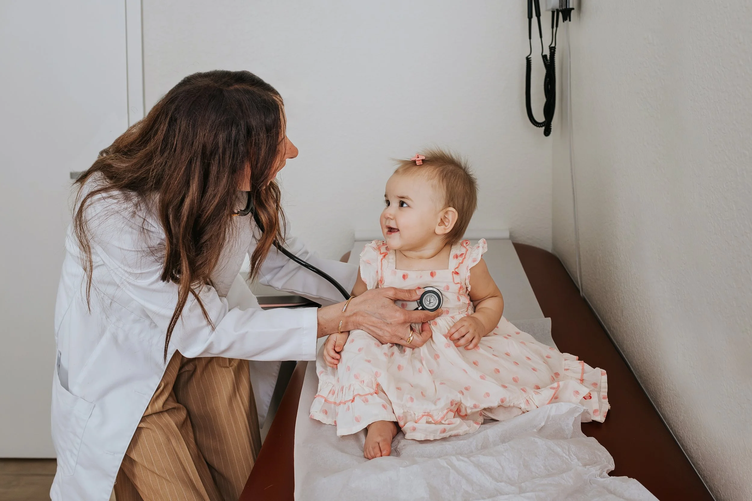 A female doctor with long brown hair examines a young girl with a stethoscope in a medical office. The girl has light brown hair with a pink bow and is wearing a pink and white dress. They are sitting on an examination table with a paper sheet, and medical equipment is visible on the wall.