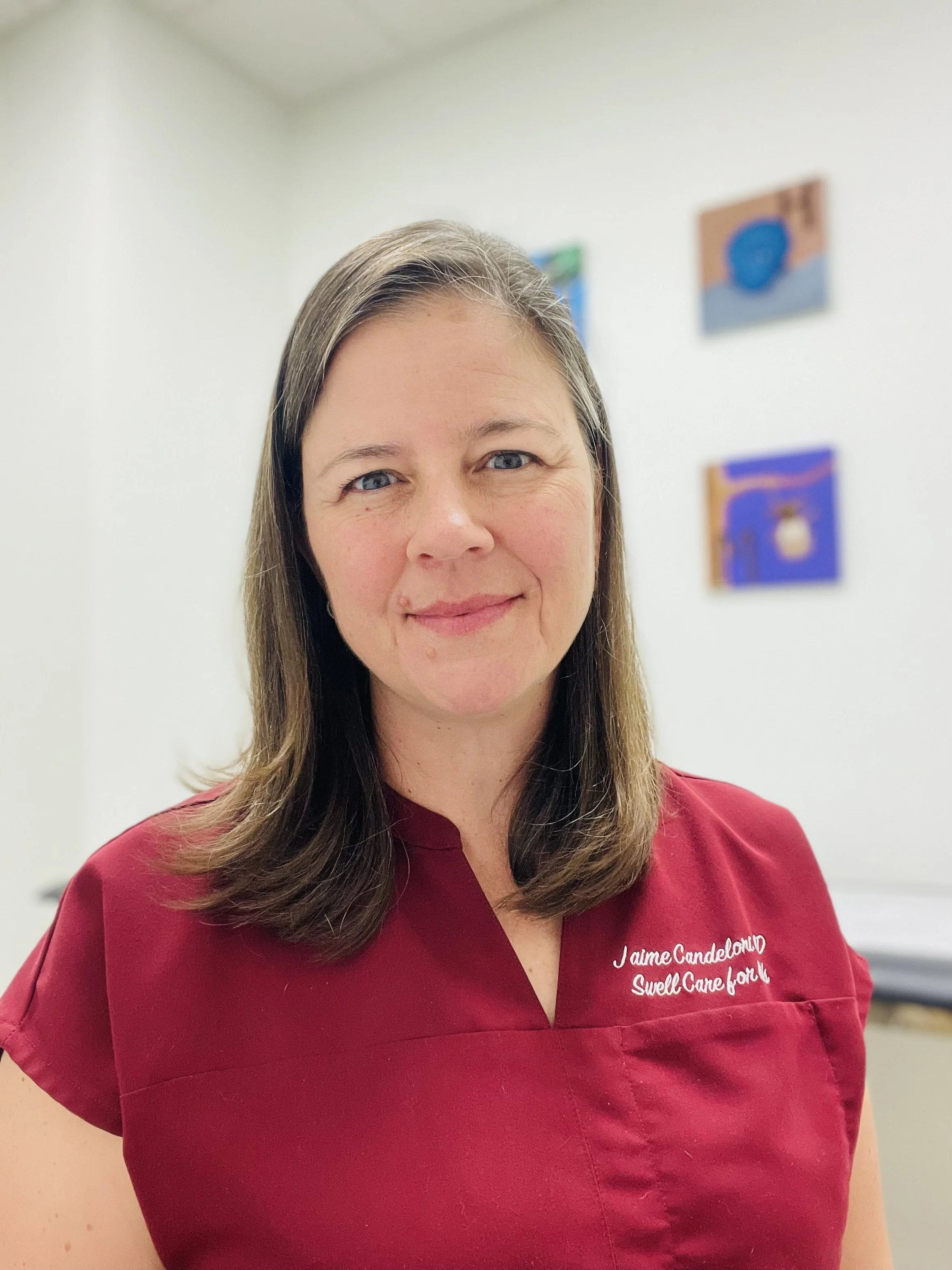 A woman with straight brown hair and blue eyes, wearing a red medical scrub top with white embroidered text that reads 'I am Candelora Swell Care for It,' standing in a clinical setting with white walls and colorful artwork in the background.