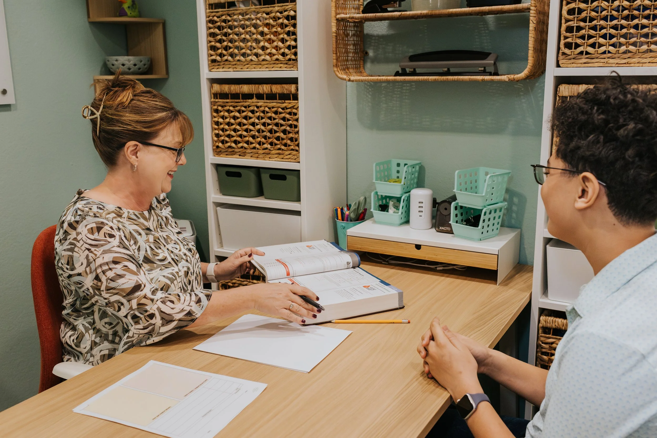 A woman and a young man sitting at a desk, engaging in a conversation, with the woman showing a pamphlet and the man listening attentively in a room with organized shelves and office supplies.
