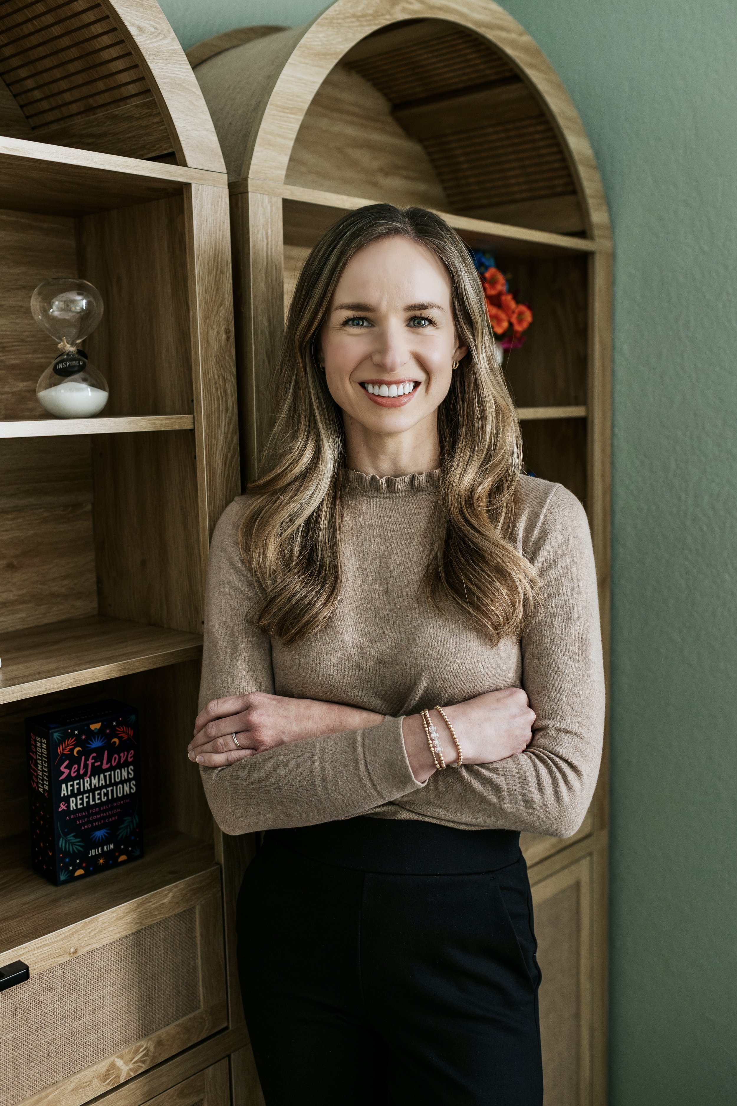 A woman with long wavy hair smiling and crossing her arms, standing in front of a wooden bookshelf with books and a small glass hourglass. She wears a beige sweater and black pants, with bracelets on her wrist, in a room with green walls.