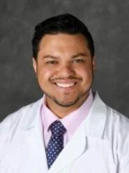 Portrait of a smiling male doctor with dark hair, wearing a white coat, a light-colored shirt, and a patterned tie, against a gray background.