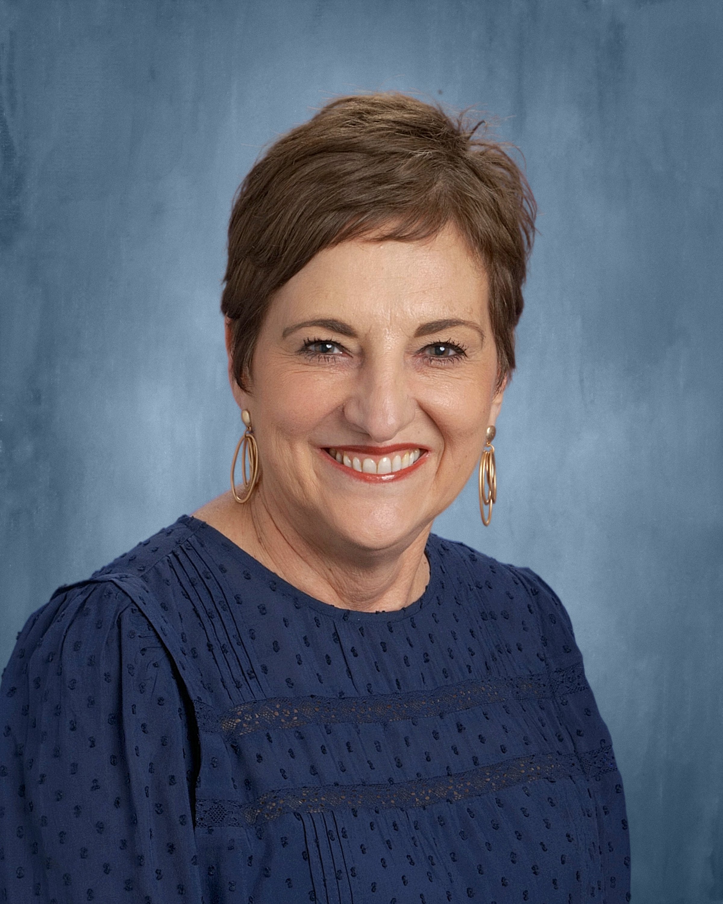 A woman smiling, wearing a navy blue dress with a textured pattern and gold earrings, standing against a blue background.