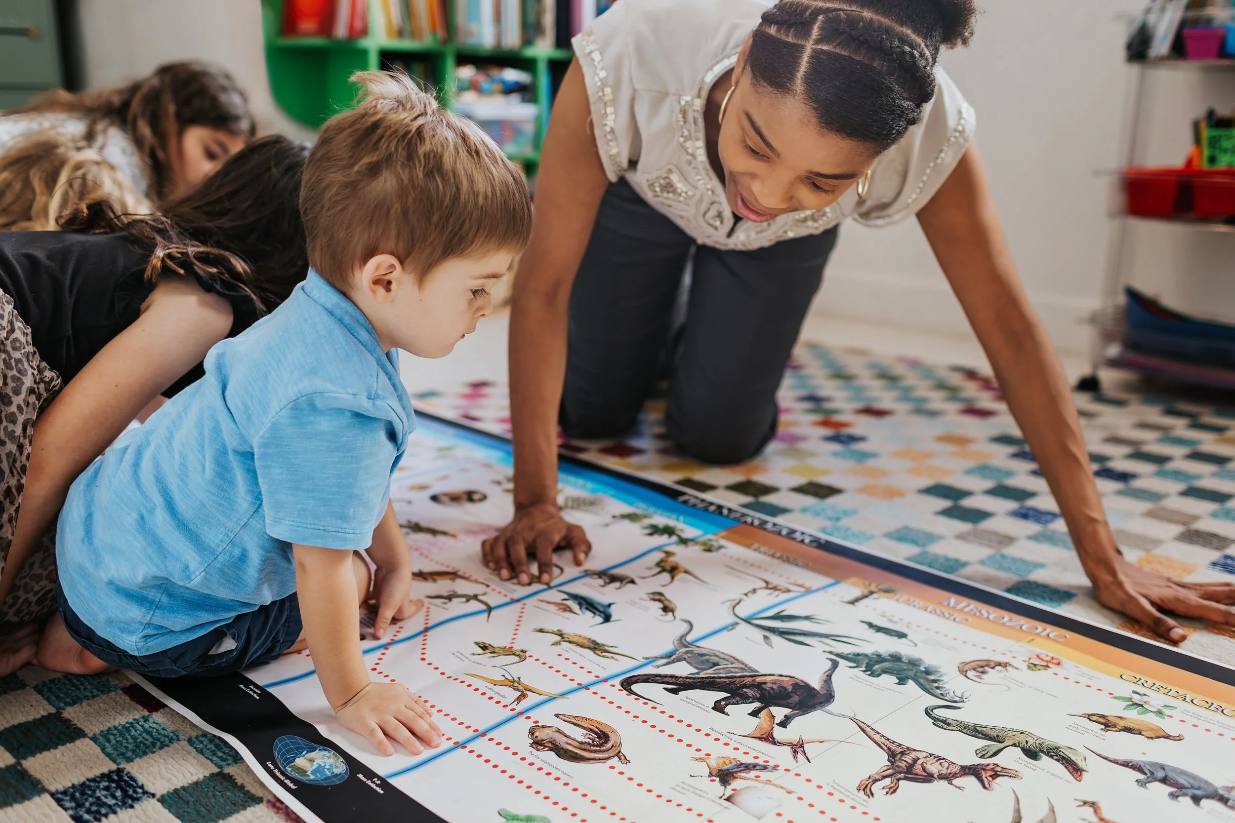 A teacher and young students examine a colorful dinosaur poster in a classroom.