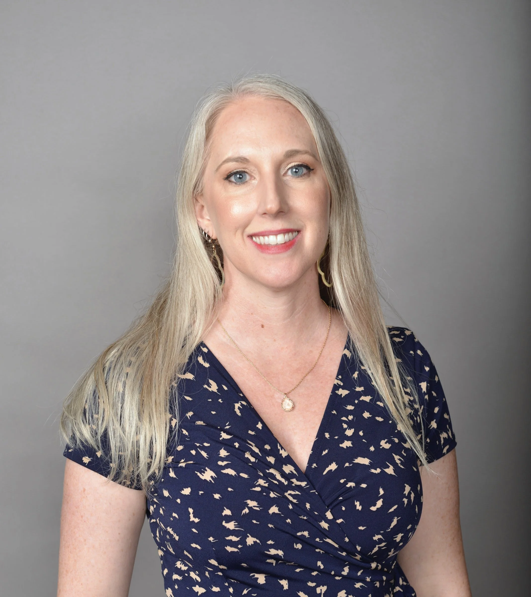 A smiling woman with long blonde hair, blue eyes, wearing a navy dress with beige animal print, gold earrings, a necklace with a pendant, and standing against a plain gray backdrop.