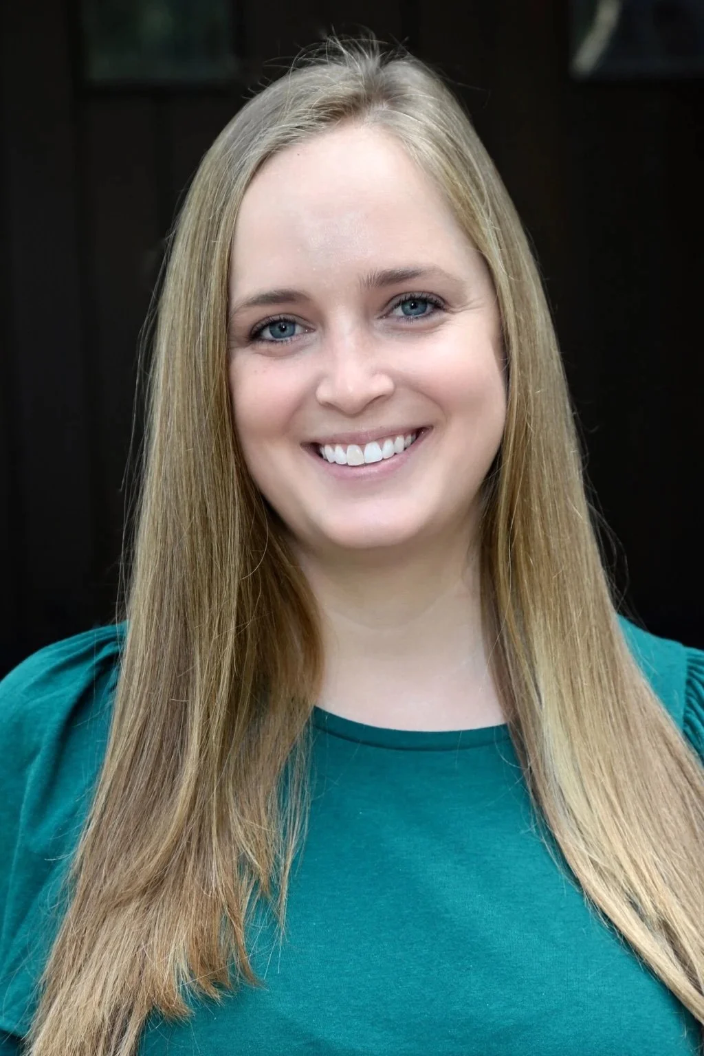 A smiling young woman with long blonde hair and blue eyes wearing a teal top, standing against a dark background.