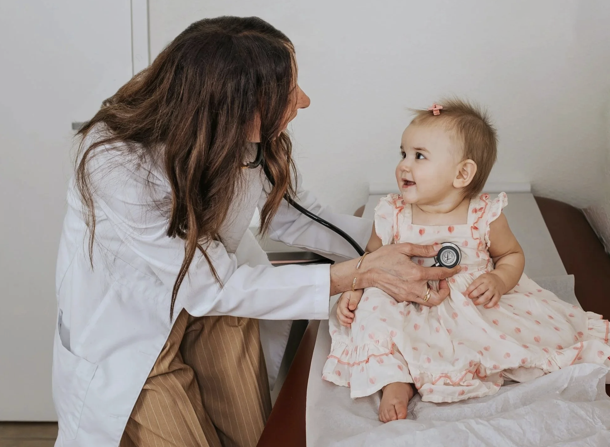 A female doctor using a stethoscope to examine a young girl who is sitting on an examination table.