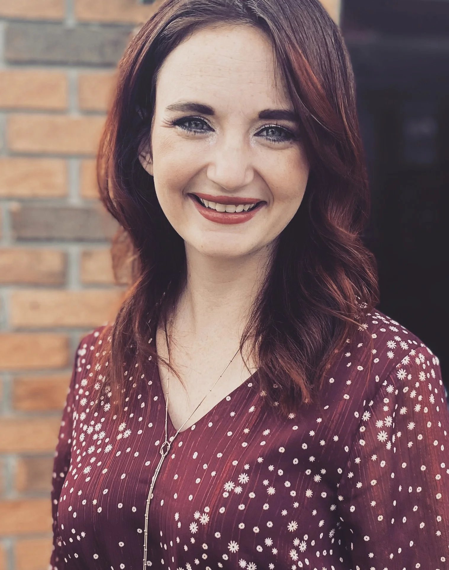 A young woman with red hair, blue eyes, and fair skin smiling, wearing a maroon dress with white floral patterns, standing in front of a brick wall.