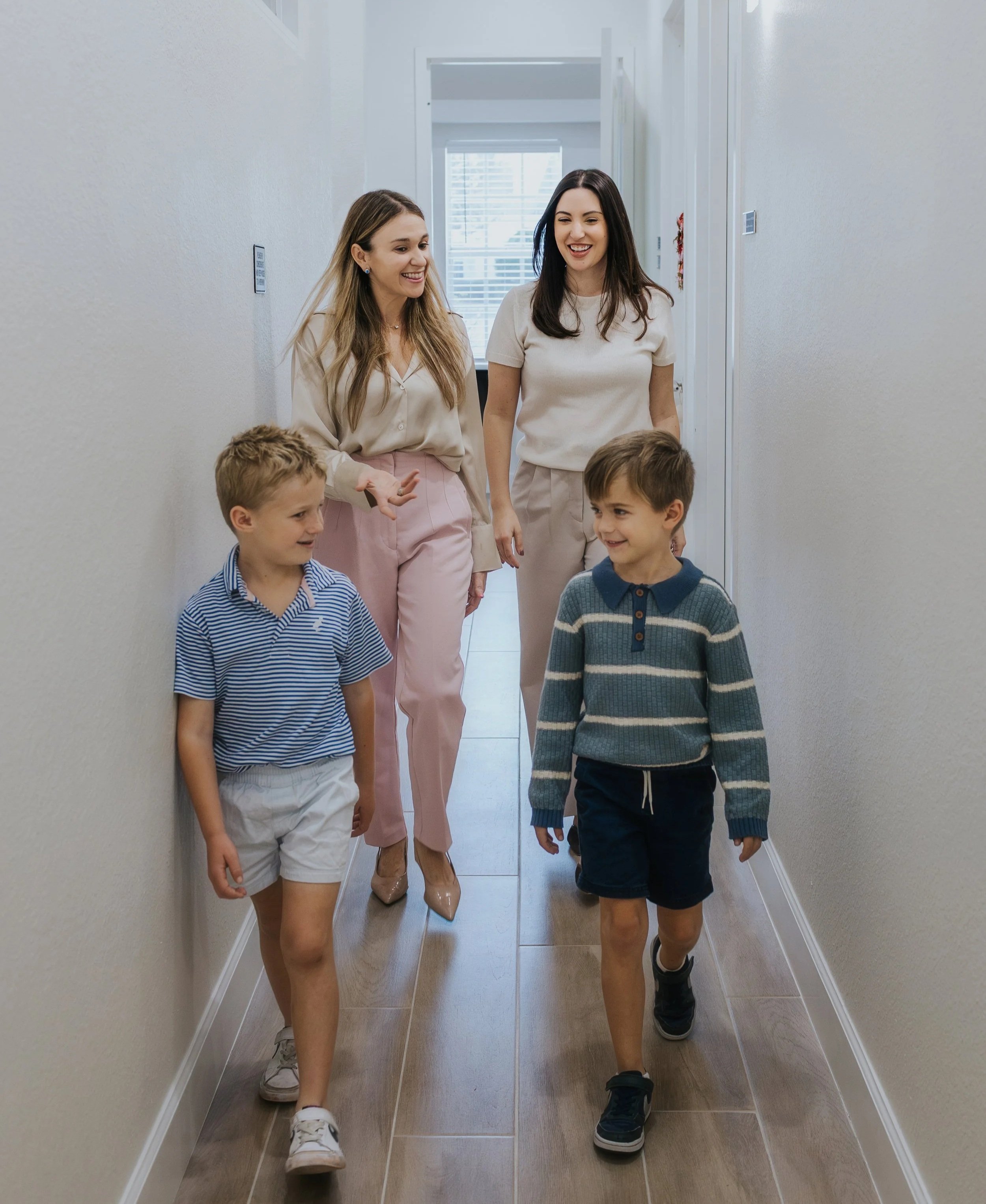 Two women with three young boys walking down a hallway, smiling and talking. The women are dressed in light-colored tops and pants, and the boys are wearing casual shirts and shorts. The hallway has beige walls and wooden floors, with a window at the end letting in natural light.
