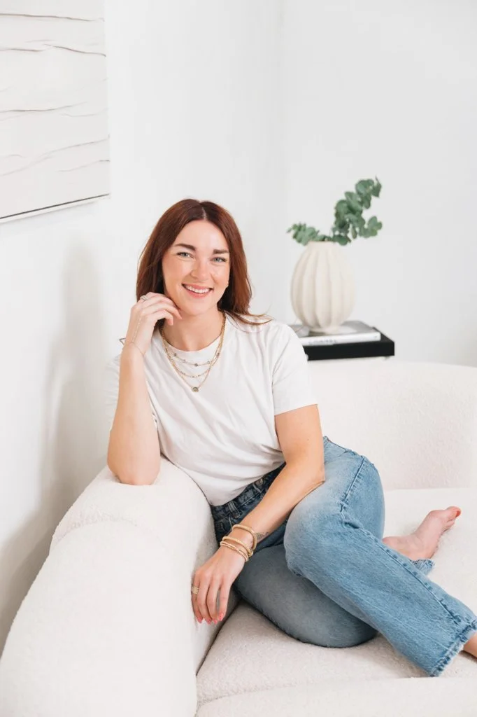 A smiling woman with shoulder-length brown hair, wearing a white t-shirt, layered necklaces, and jeans, sitting on a white sofa in a bright, minimalistic room with a white wall and a decorative vase with green foliage in the background.