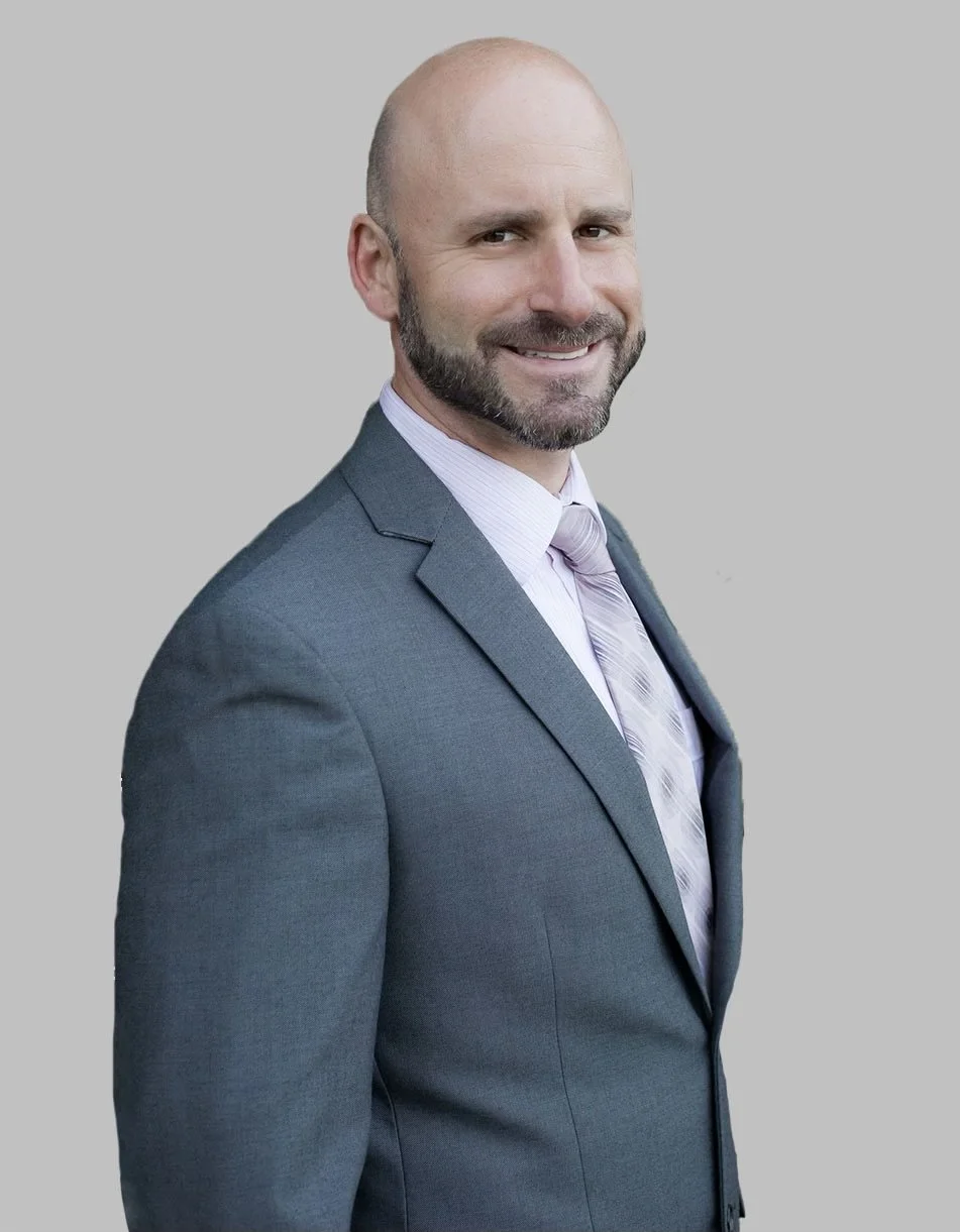 A bald man with a beard wearing a gray suit, white shirt, and patterned tie, smiling against a plain gray background.