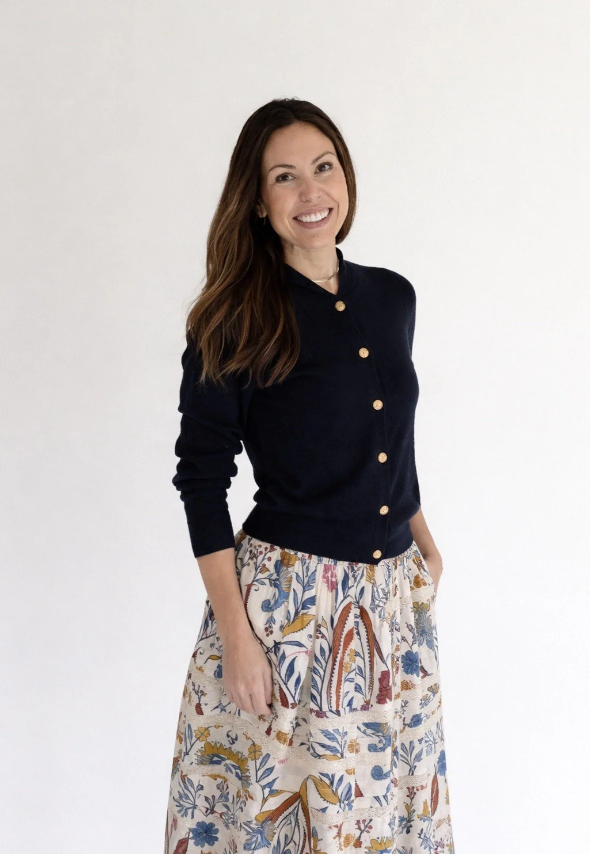 A woman with long brown hair smiling, wearing a black cardigan with gold buttons and a beige skirt with a floral pattern, standing against a plain white wall.