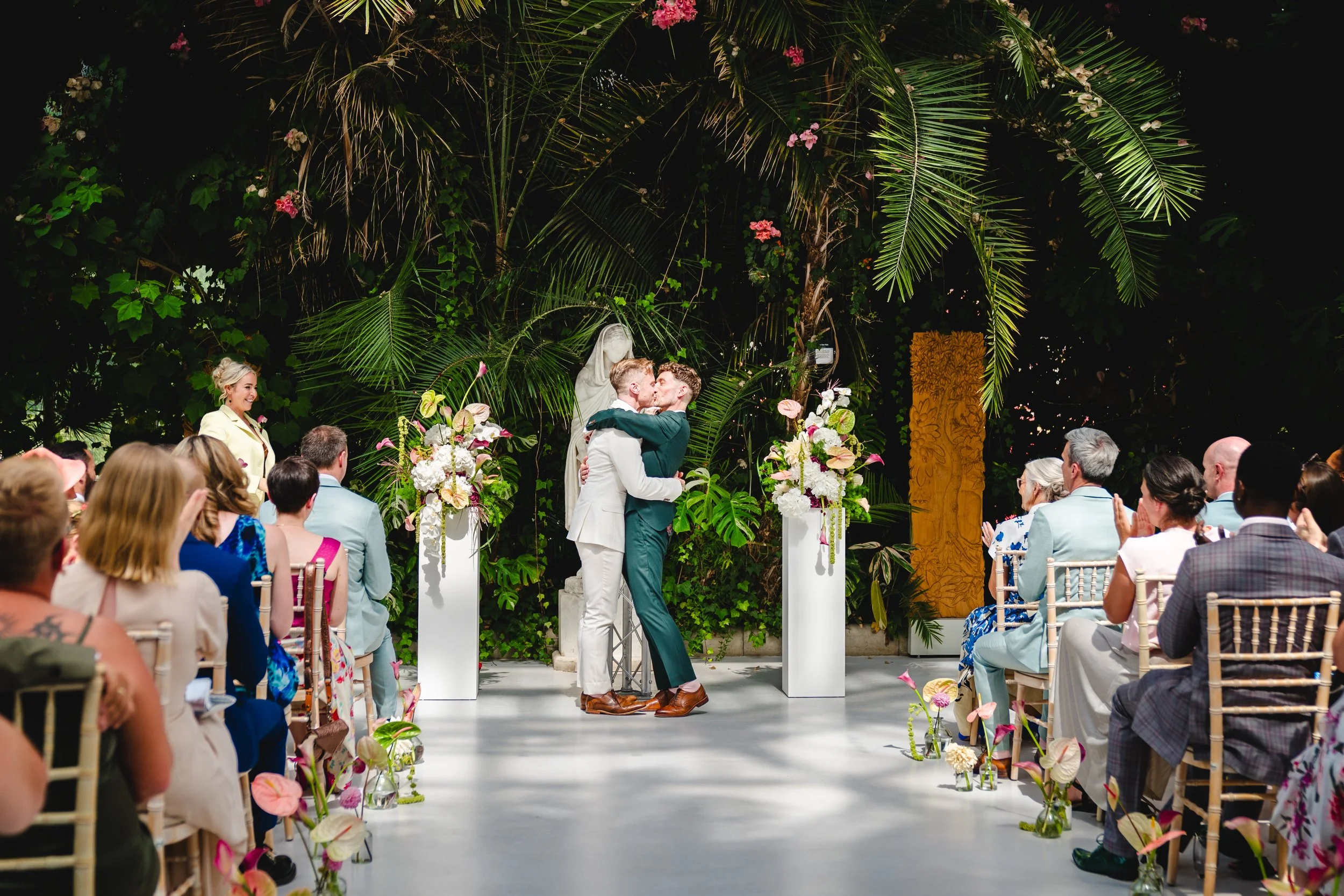 two men have their first kiss on their wedding day in a tropical setting full of plants. the LGBT wedding celebrant, Philli Boyle, your celebrant looks on wearing a yellow suit and smiling