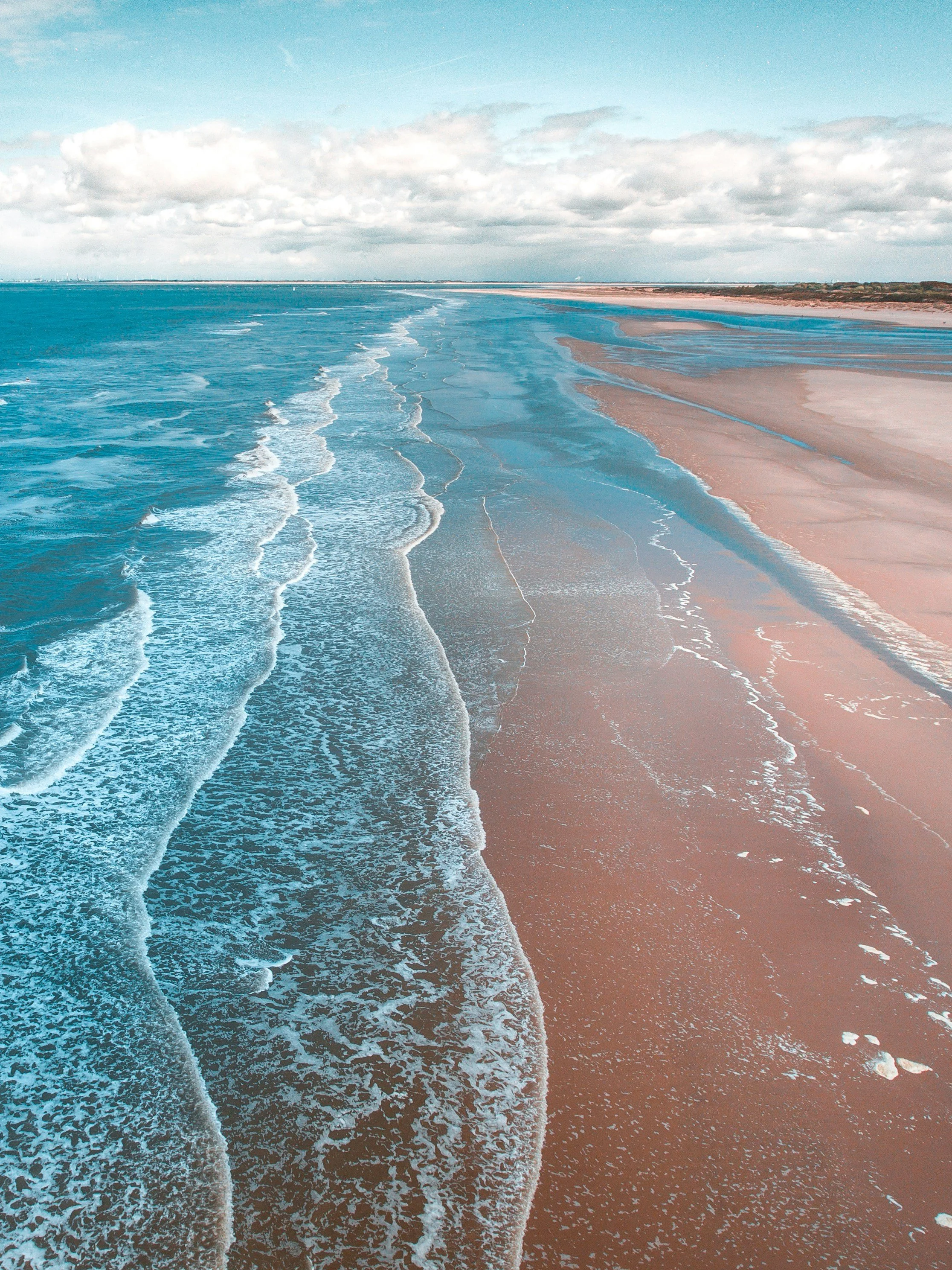 Aerial view of a beach with blue ocean waves and sandy shore, under a partly cloudy sky.