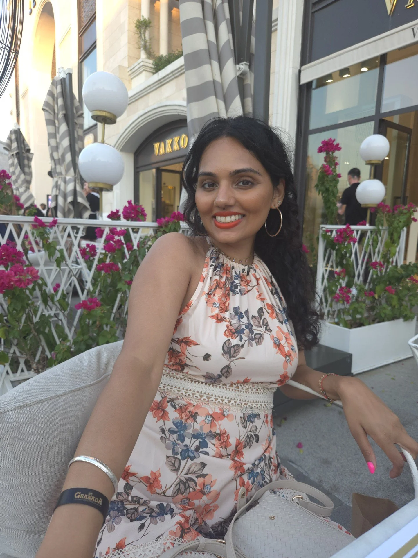 A woman with dark curly hair smiling at a table outdoors at a cafe with pink flowers and striped awnings.