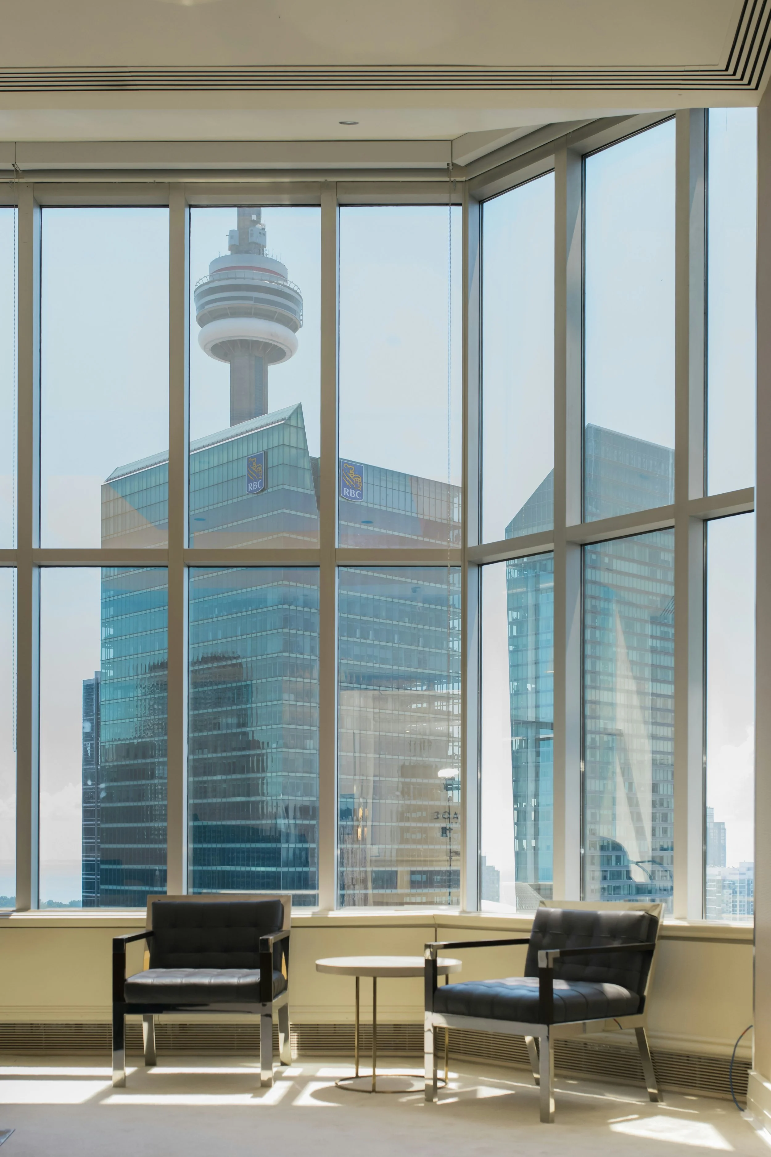 Interior of a modern office with two black armchairs and a small round table, large windows with a city view of tall buildings and a tower in the background.