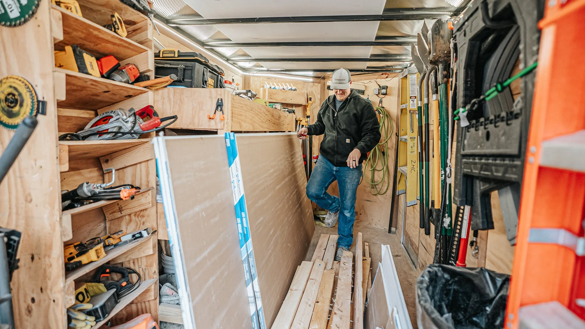 A man in a black hoodie and blue jeans in a workshop, arranging wood panels among various tools on shelves and hanging on the walls.